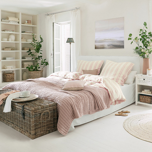 Camera accogliente con biancheria da letto bianca e coperta rosa, decorazioni naturali e baule in vimini
