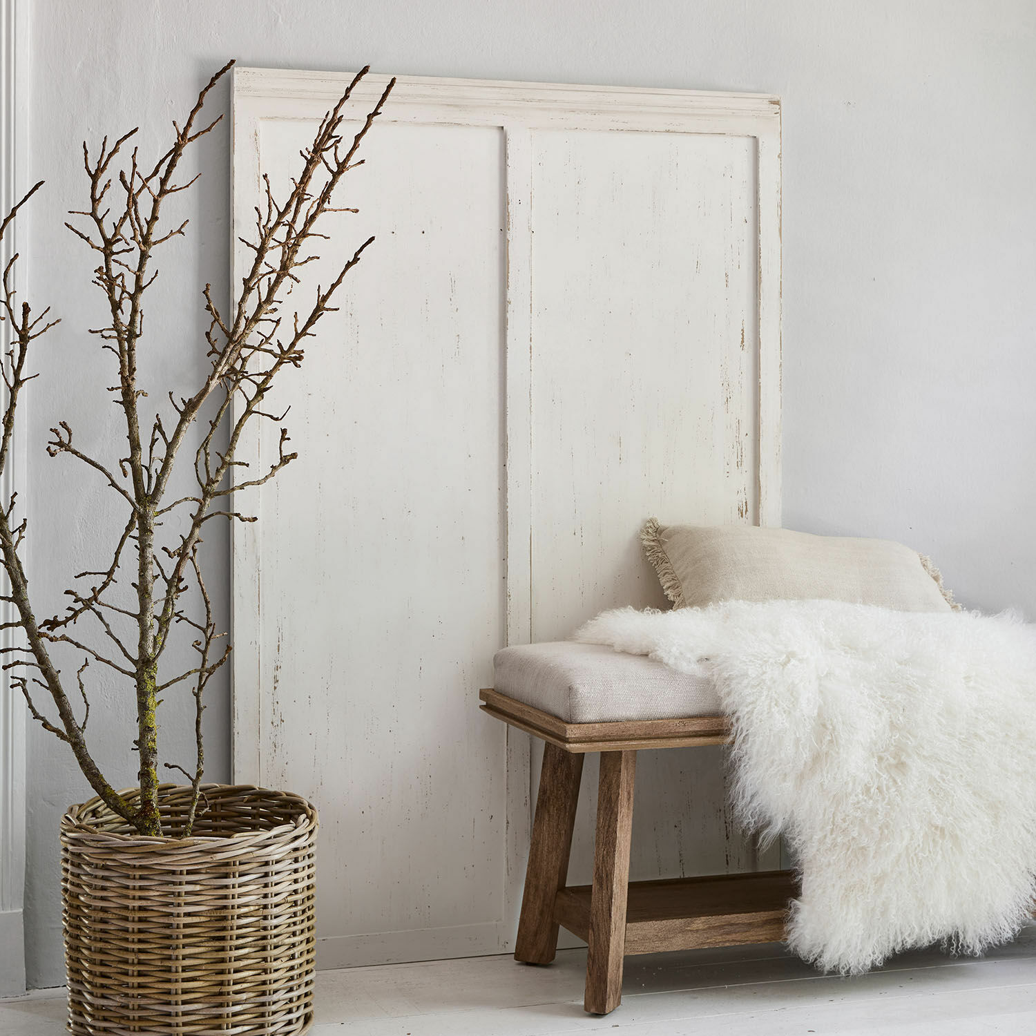 Pannello Evanston Panca in legno con cuscino beige e coperta bianca di pelliccia accanto a un cesto di vimini con rami