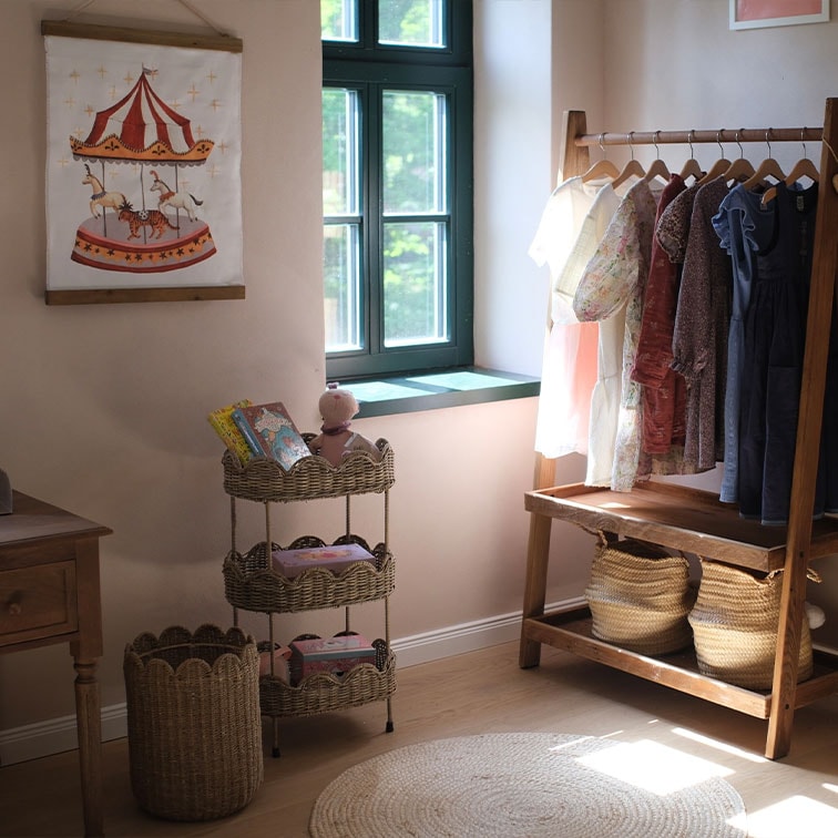 Scaffale in vimini a tre livelli con giocattoli e abbigliamento per bambini su appendiabiti in legno in stanza luminosa