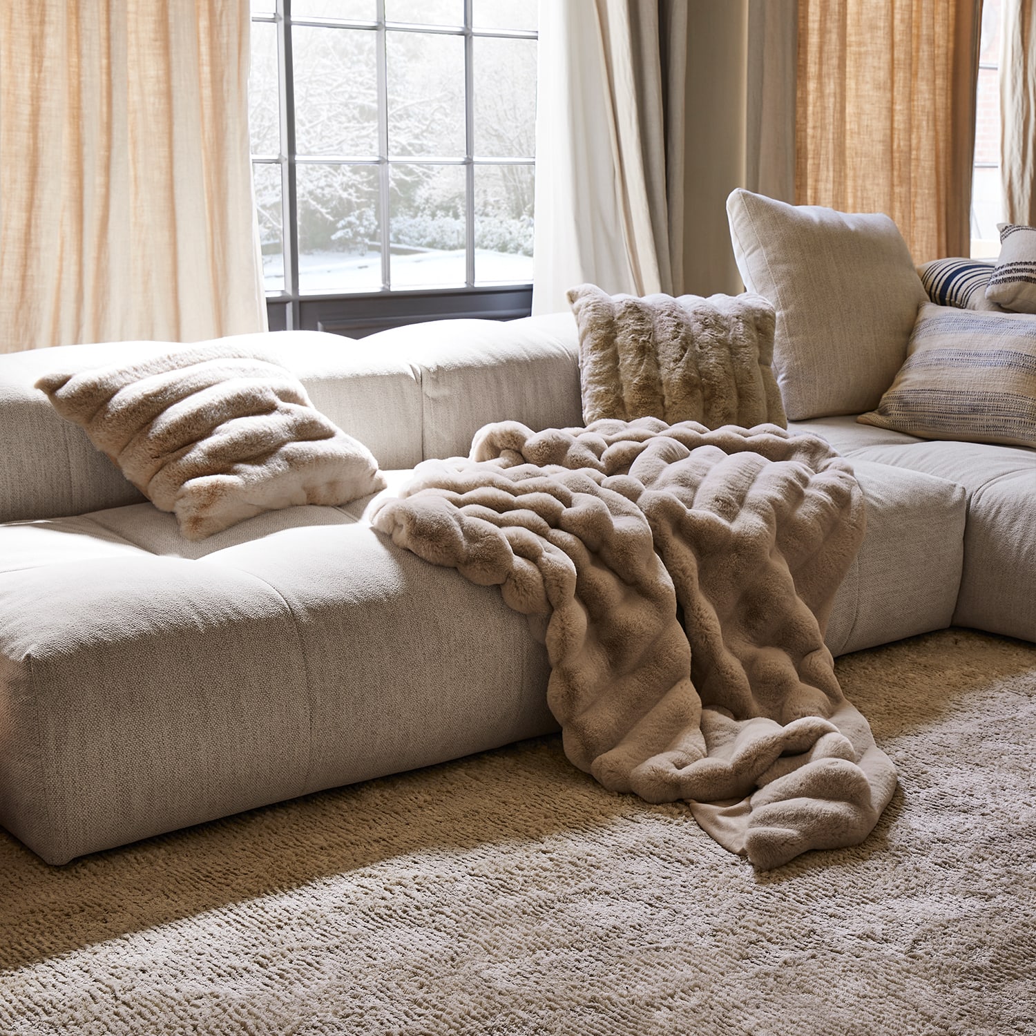 Coperta e cuscini in pelliccia sintetica beige su divano chiaro in soggiorno accogliente con vista su giardino innevato