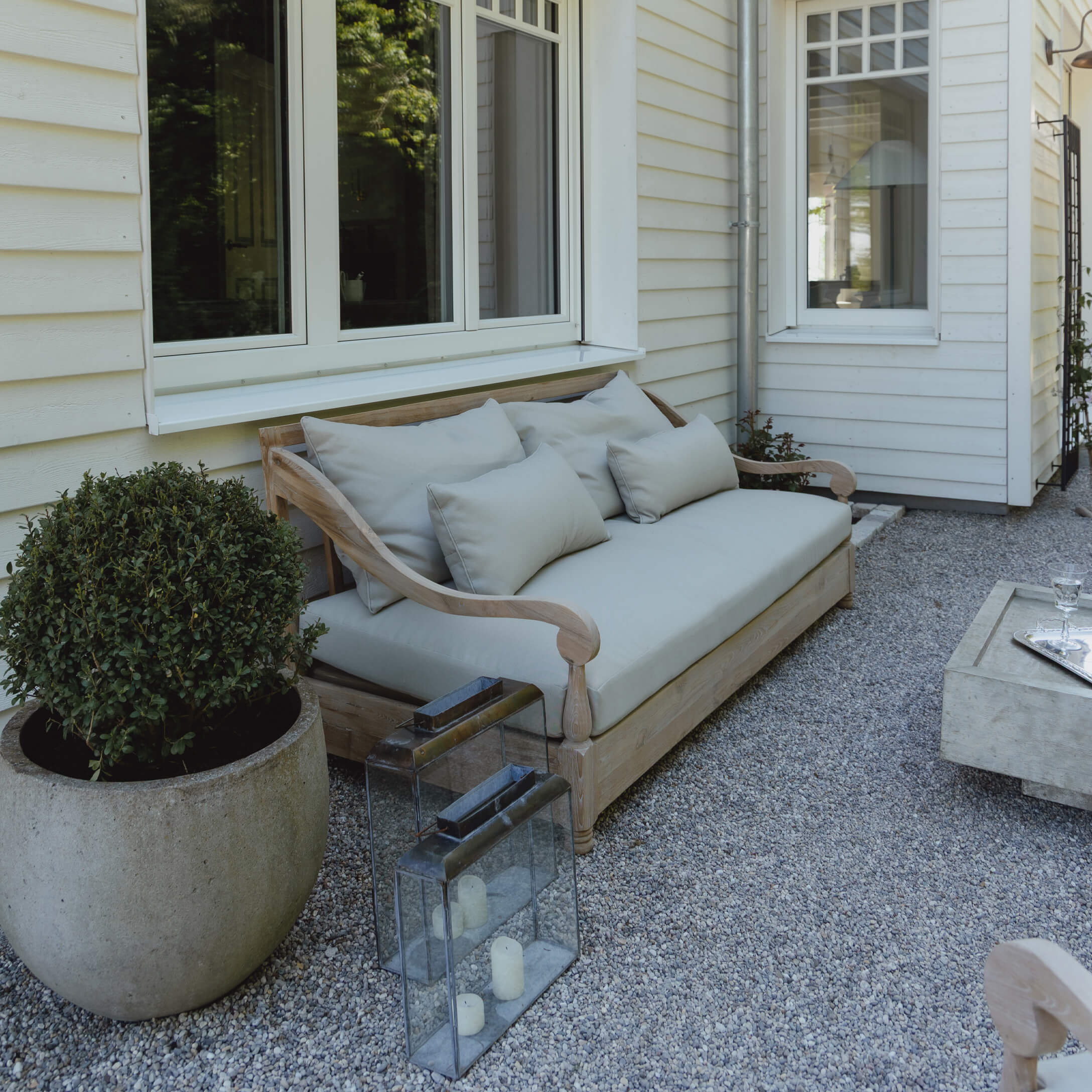 Divano da giardino in legno con cuscini grigio chiaro su patio ghiaioso davanti a casa bianca