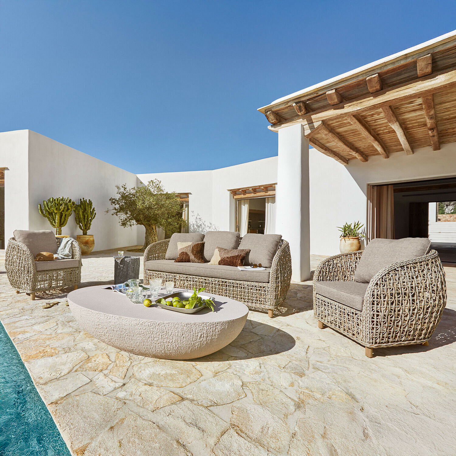 Set di mobili da giardino in rattan beige con cuscini su terrazza in pietra vicino a piscina soleggiata