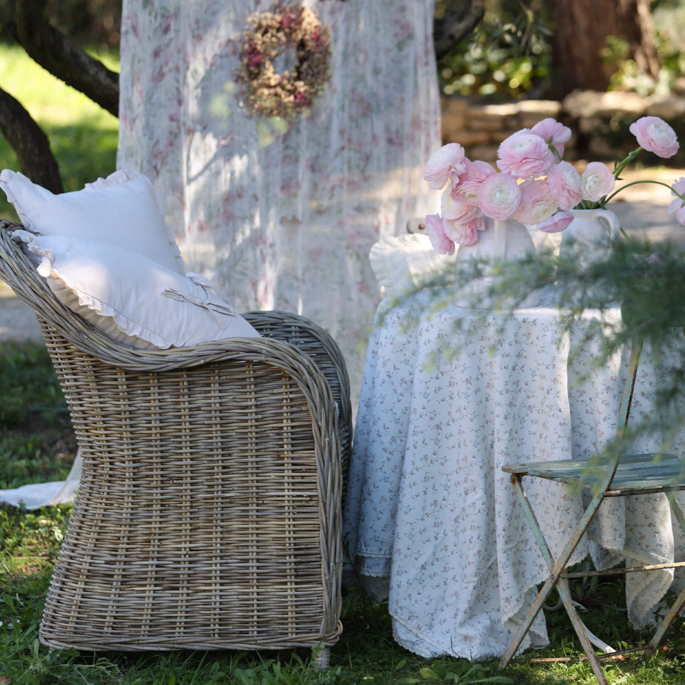 Poltrona in vimini con cuscini bianchi accanto a tavolo con tovaglia floreale e fiori rosa in giardino