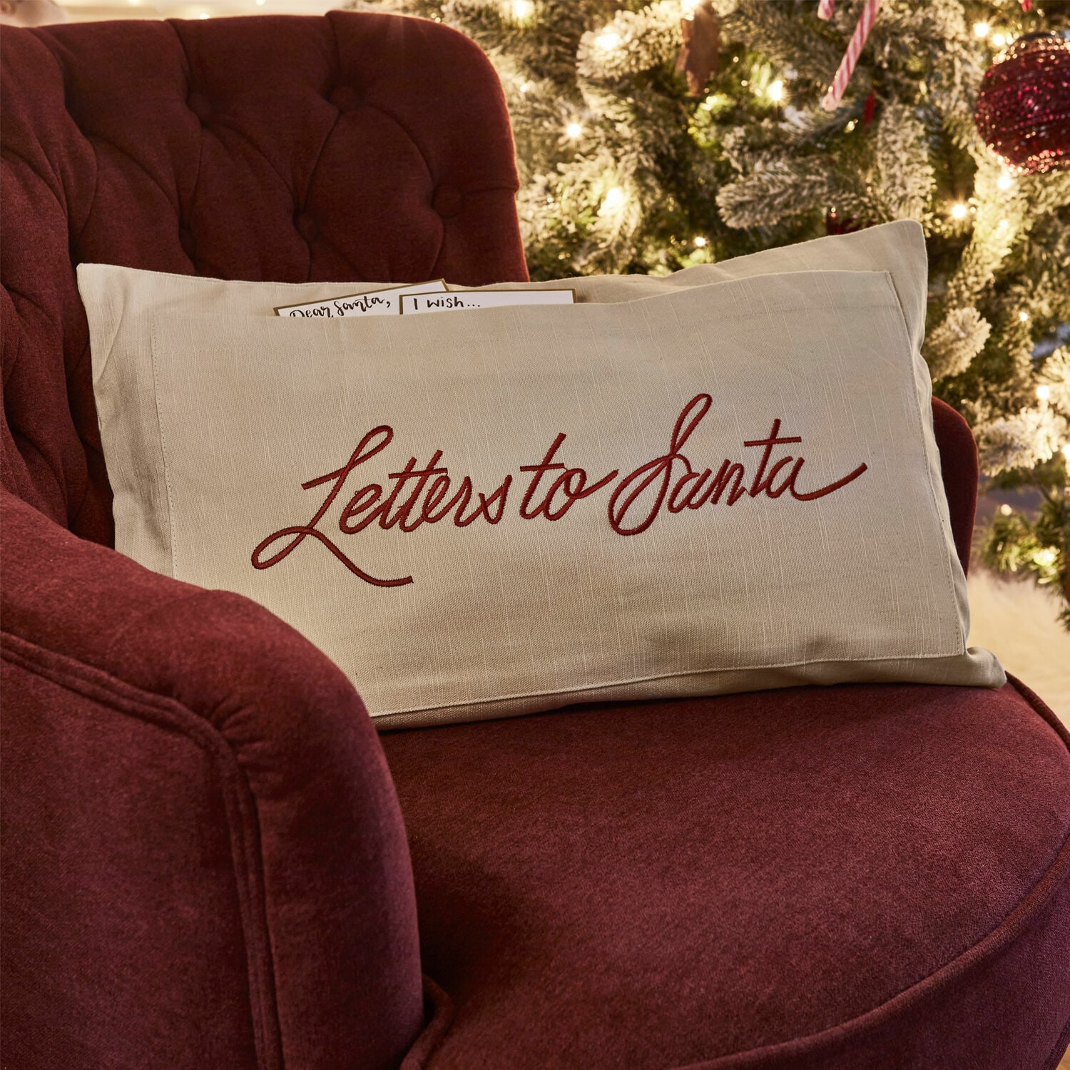 Copricuscino Welenzas Cuscino beige in lino con scritta rossa Letters to Santa su poltrona bordeaux vicino all'albero di Natale