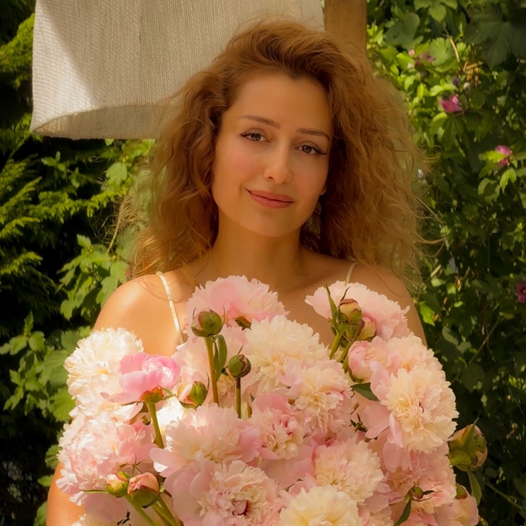 Donna con capelli lunghi che tiene un grande mazzo di peonie rosa davanti a un giardino verde
