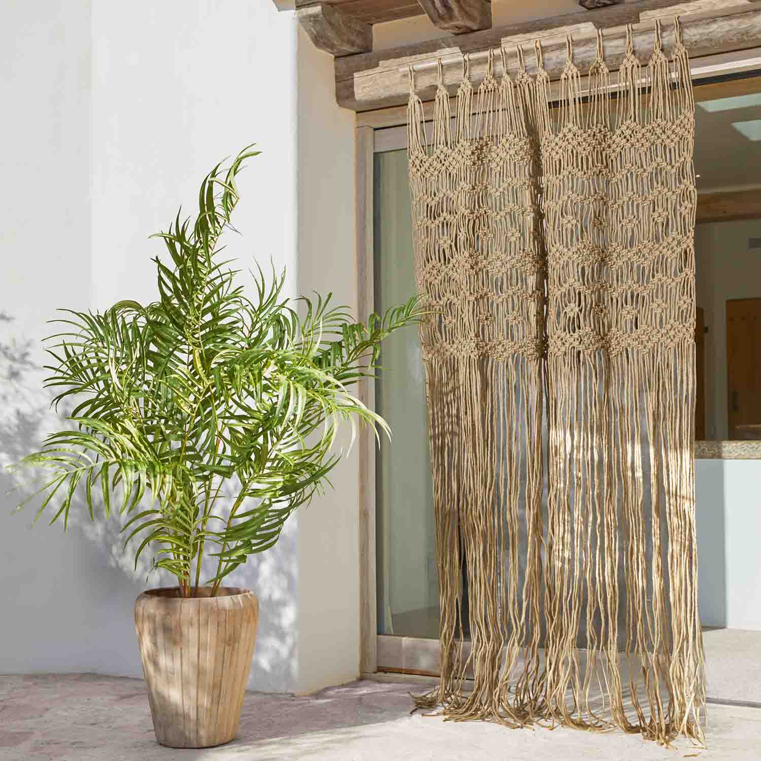 Tenda macramè beige in cotone con lunghe frange davanti a porta finestra, pianta in vaso accanto
