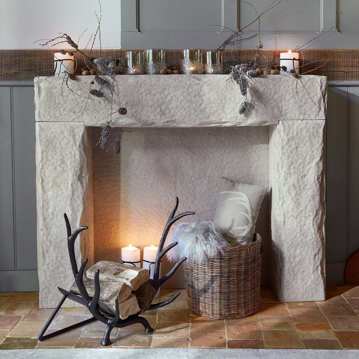 Camino in pietra con rami decorativi, candele e cesta in vimini con cuscino e coperta in pelliccia sintetica