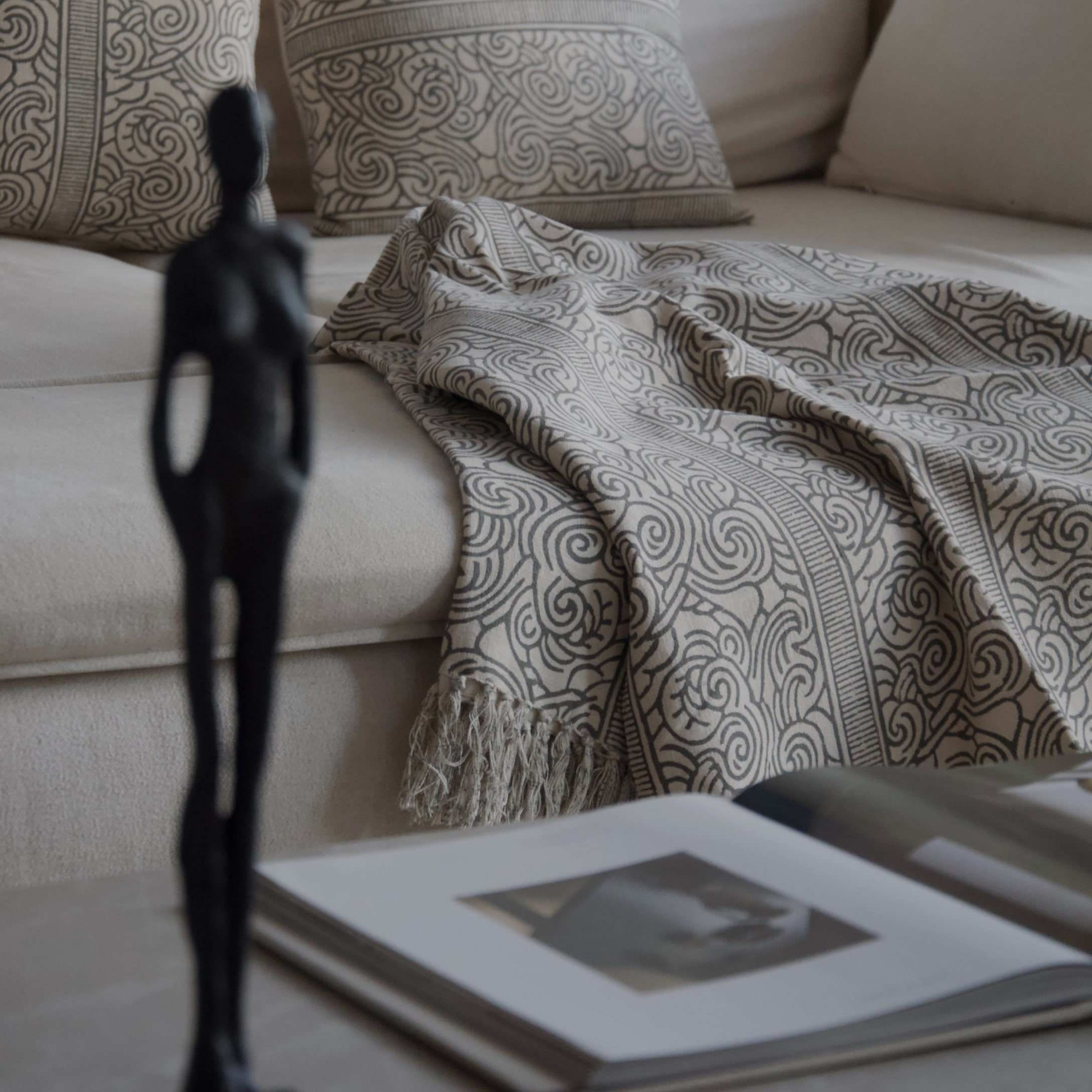 Coperta beige con motivo grigio e frange su divano chiaro con cuscini decorativi e libro aperto