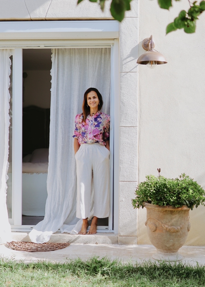 Donna con camicetta floreale e pantaloni bianchi in porta con tenda bianca e vaso con pianta verde