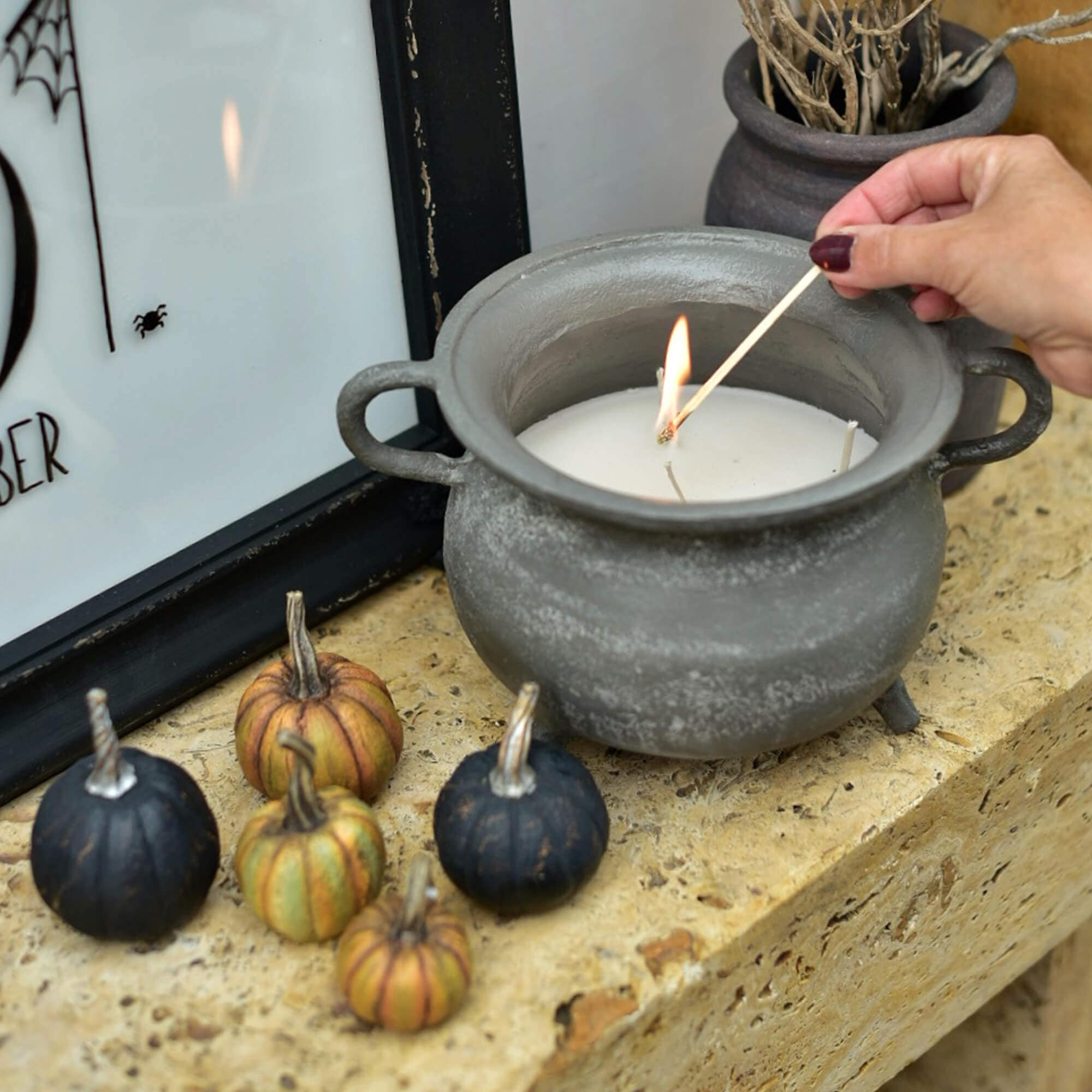Candela in ceramica grigia a forma di calderone con cera bianca e mini zucche decorative su mensola in pietra