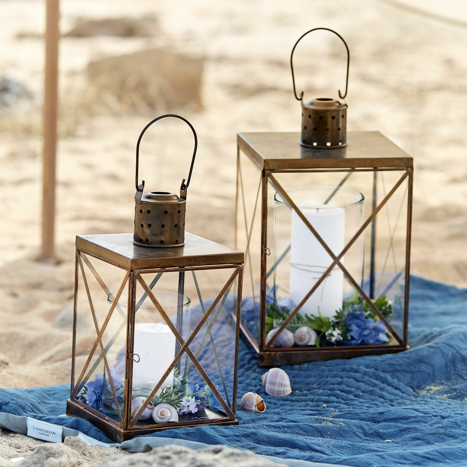 Coppia di lanterne Garance Due lanterne in metallo marrone con candele bianche e decorazioni su coperta blu in spiaggia