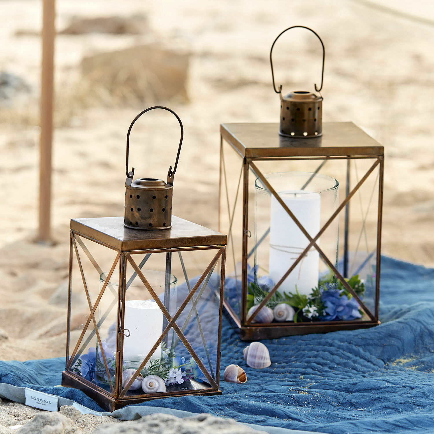 Coppia di lanterne Garance Due lanterne in metallo marrone con candele bianche e decorazioni su coperta blu in spiaggia
