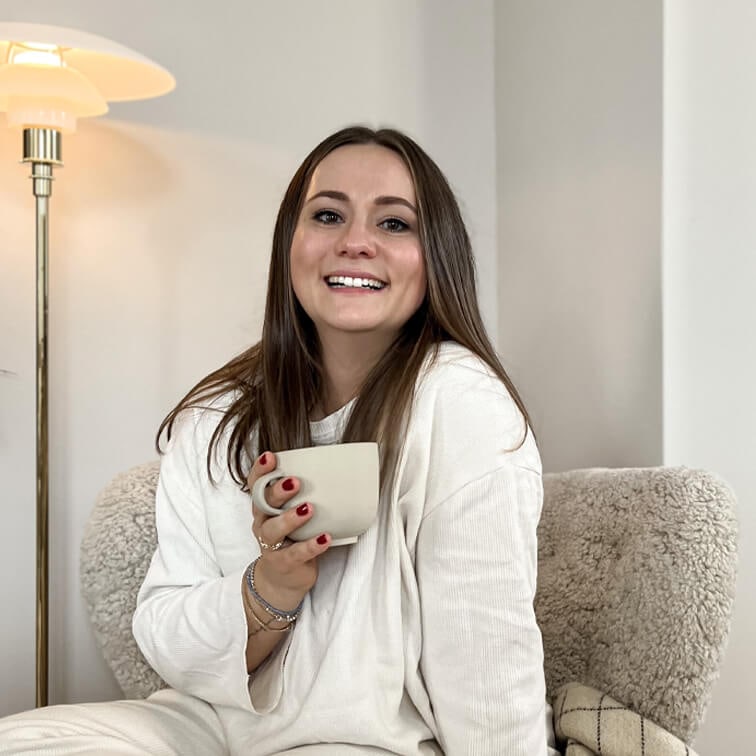 Donna sorridente con maglione bianco che tiene una tazza beige seduta su una poltrona morbida in un interno accogliente