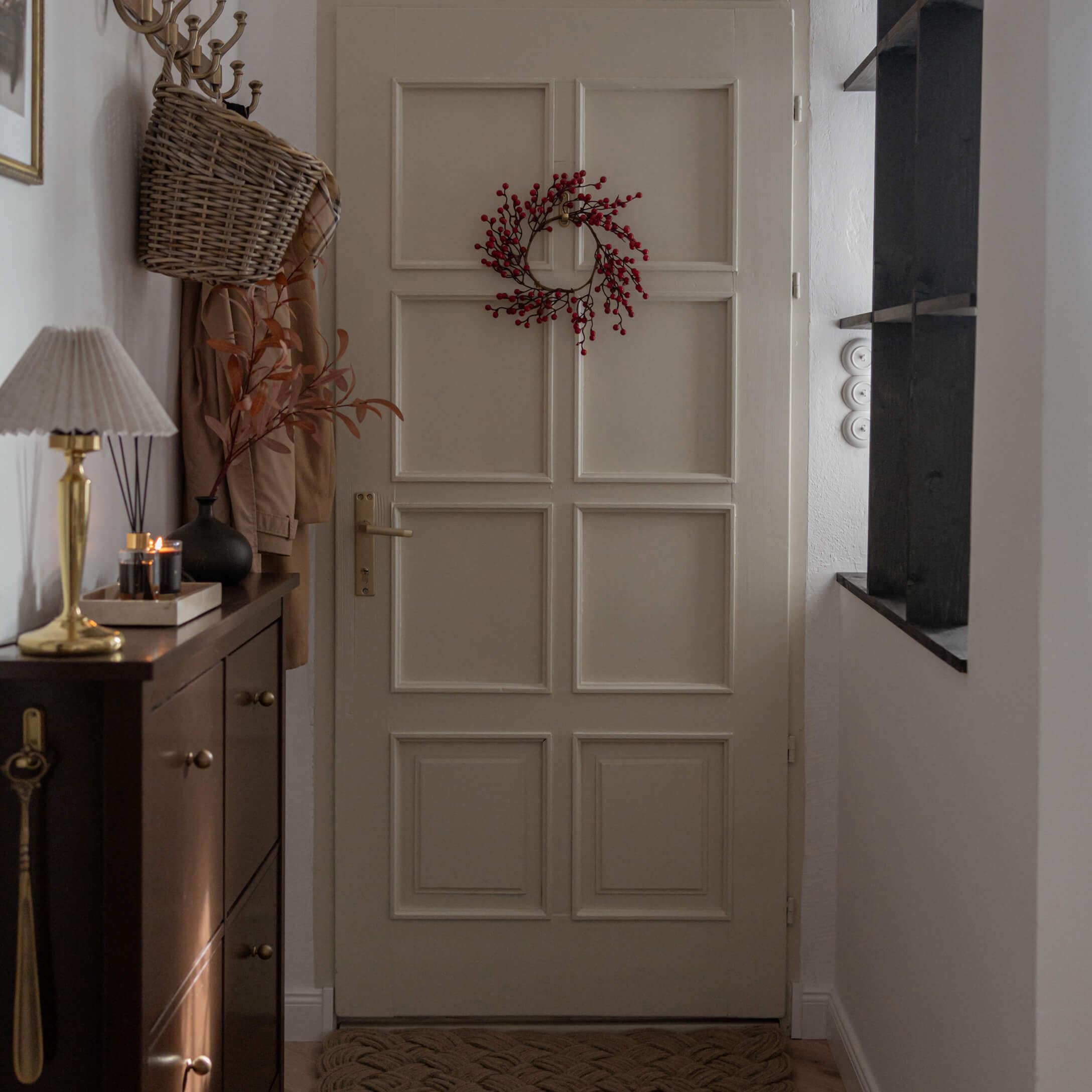 Ingresso con porta beige decorata con corona rossa e mobile scuro con lampada e candele accese