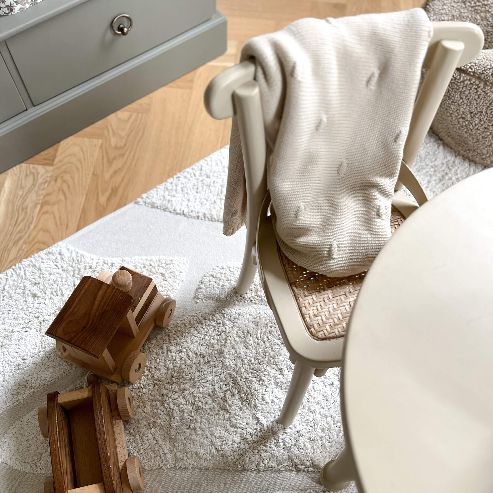Coperta beige lavorata a maglia su sedia con seduta intrecciata accanto a giocattoli in legno su tappeto chiaro