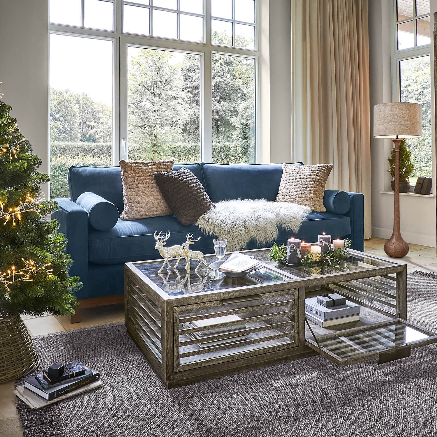 Soggiorno accogliente con divano blu scuro, tavolino in vetro e decorazioni natalizie con albero e candele