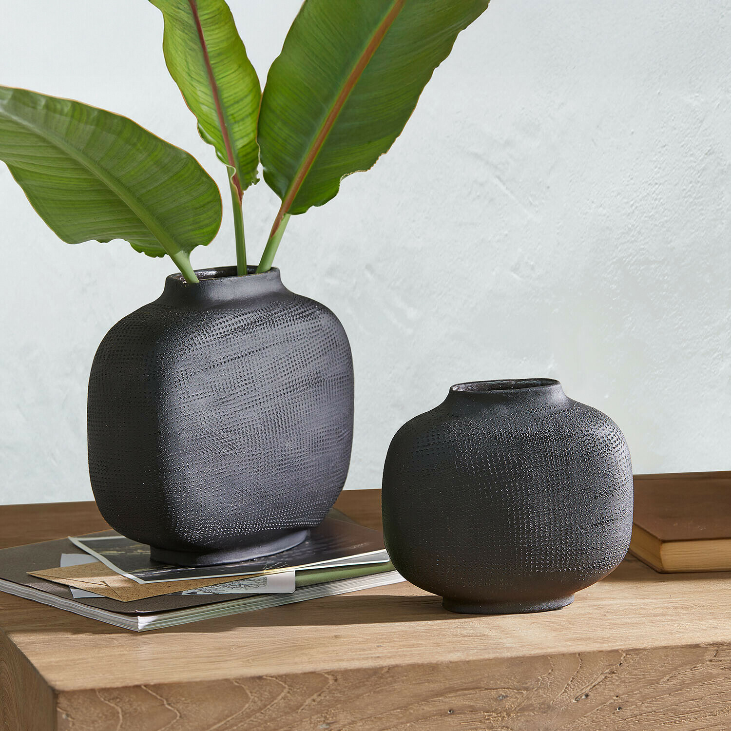 Coppia di vasi Fjordis Due vasi in ceramica nera opaca con texture su tavolo in legno con foglie verdi e libri