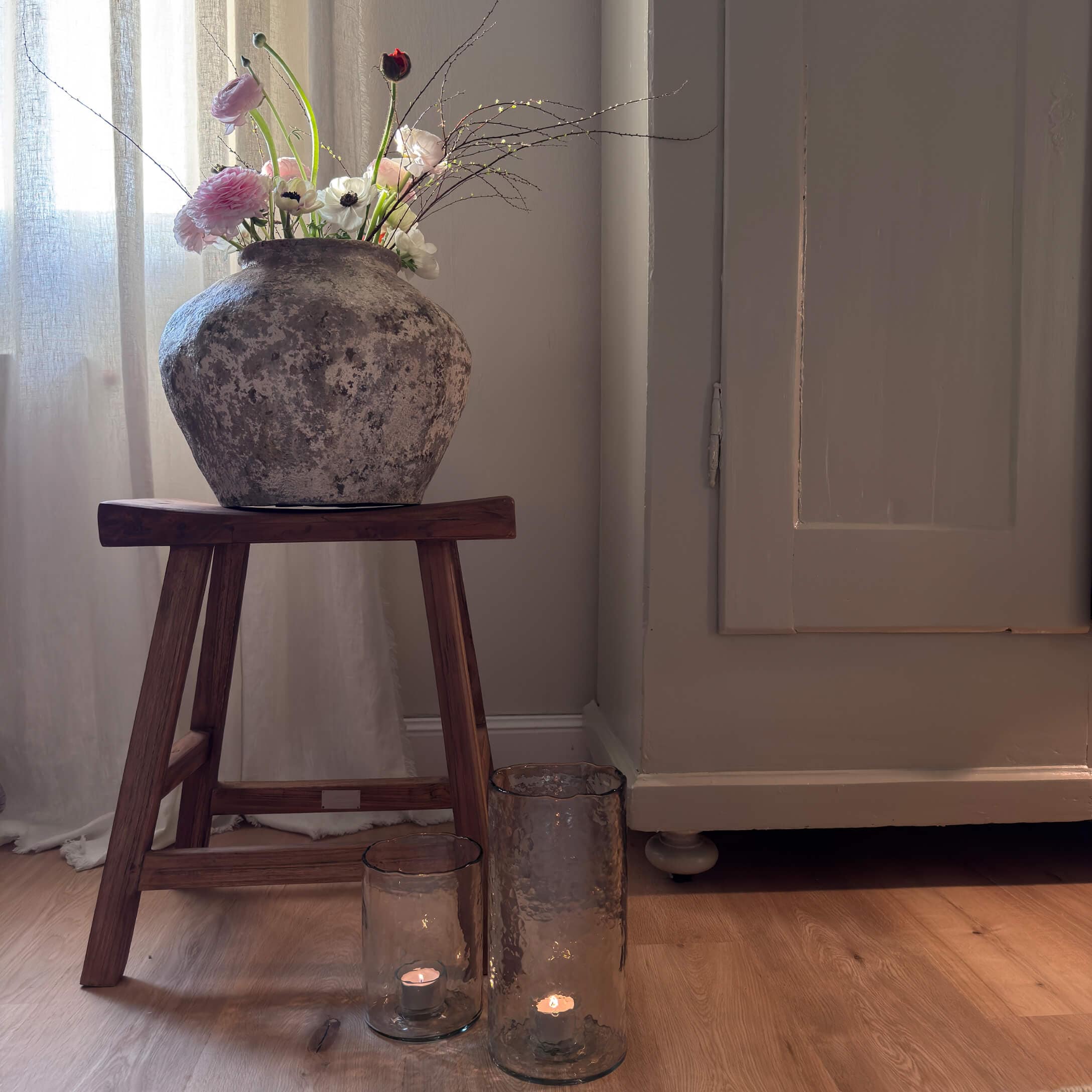 Vaso rustico in pietra con fiori su sgabello in legno accanto a candele in lanterne di vetro