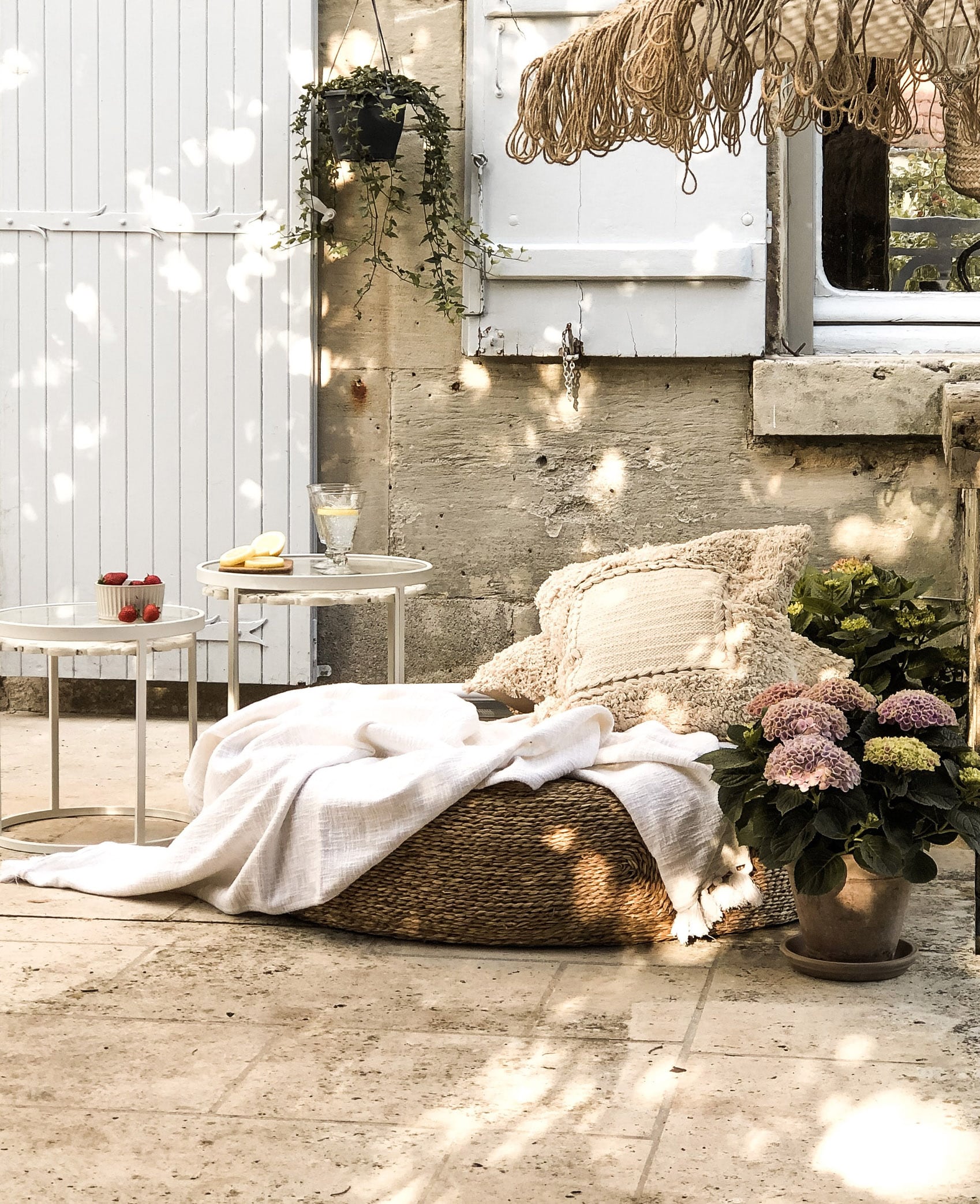Angolo accogliente con pouf in vimini, coperta crema e cuscino su terrazza in pietra con fiori in vaso.