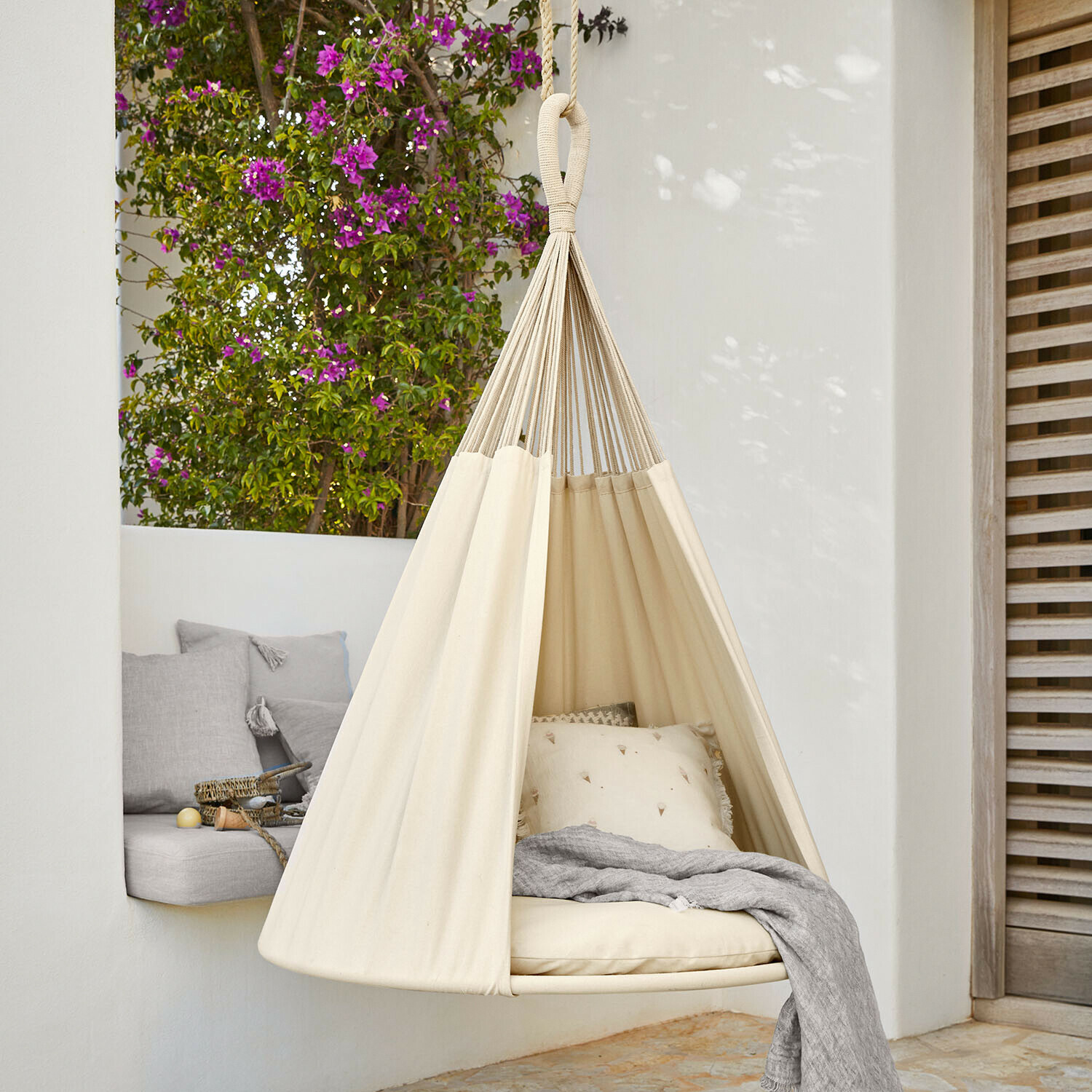 Amaca sospesa beige in cotone con cuscini e coperta grigia su terrazza fiorita