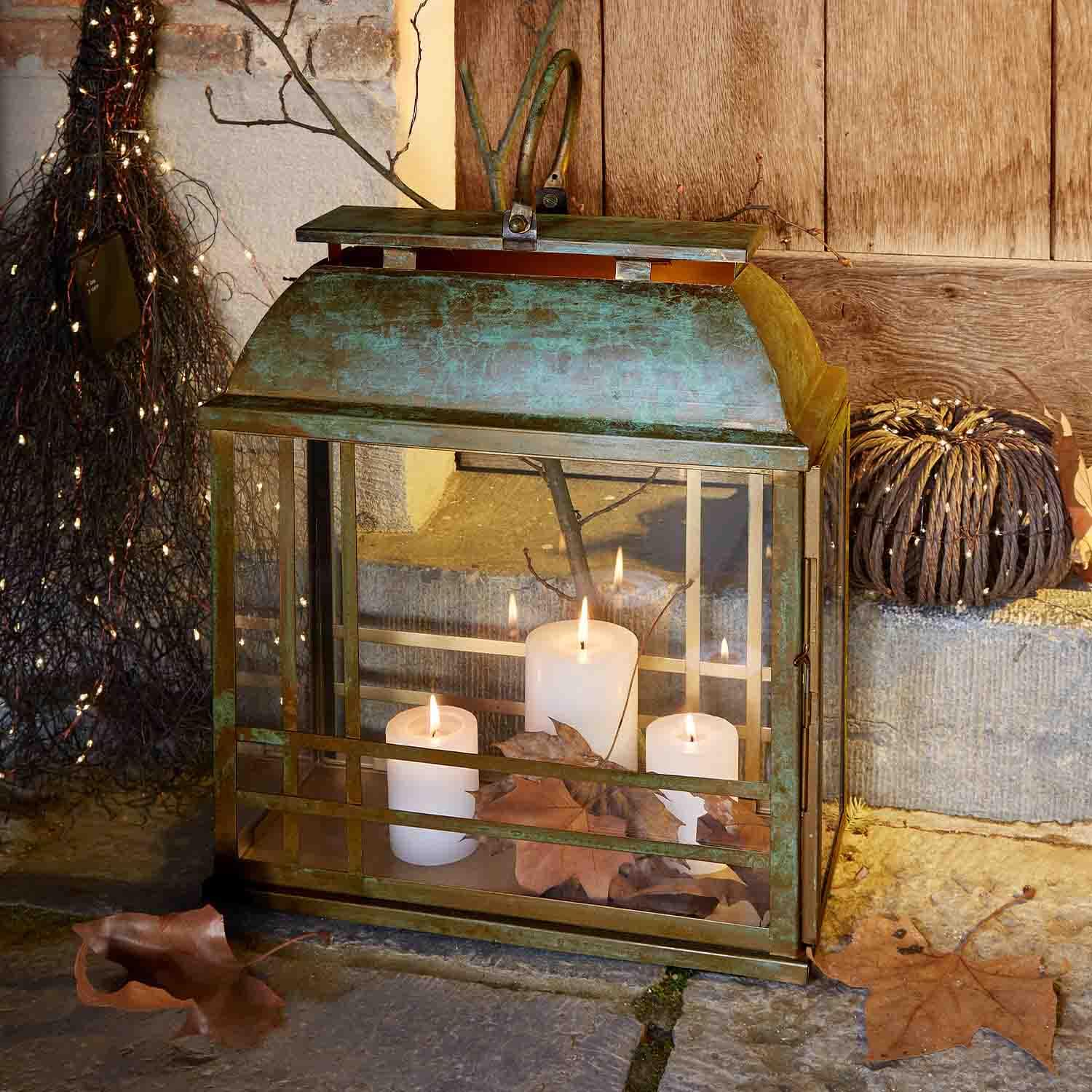 Lanterna in metallo con finitura a patina, tre candele bianche e foglie secche su pavimento in pietra