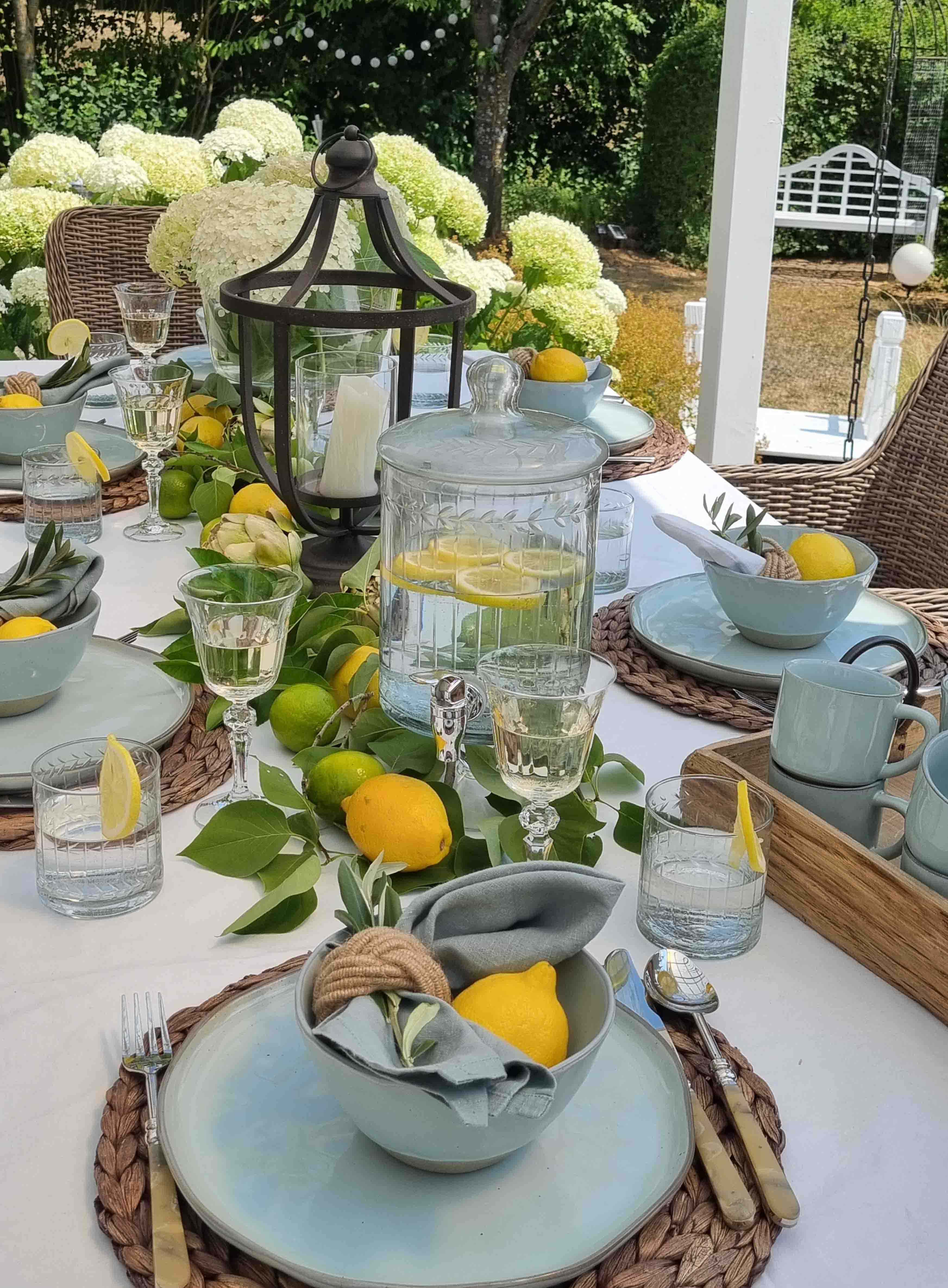 Tavolo estivo da giardino con ceramica azzurra, limoni e lanterna, fiori bianchi sullo sfondo
