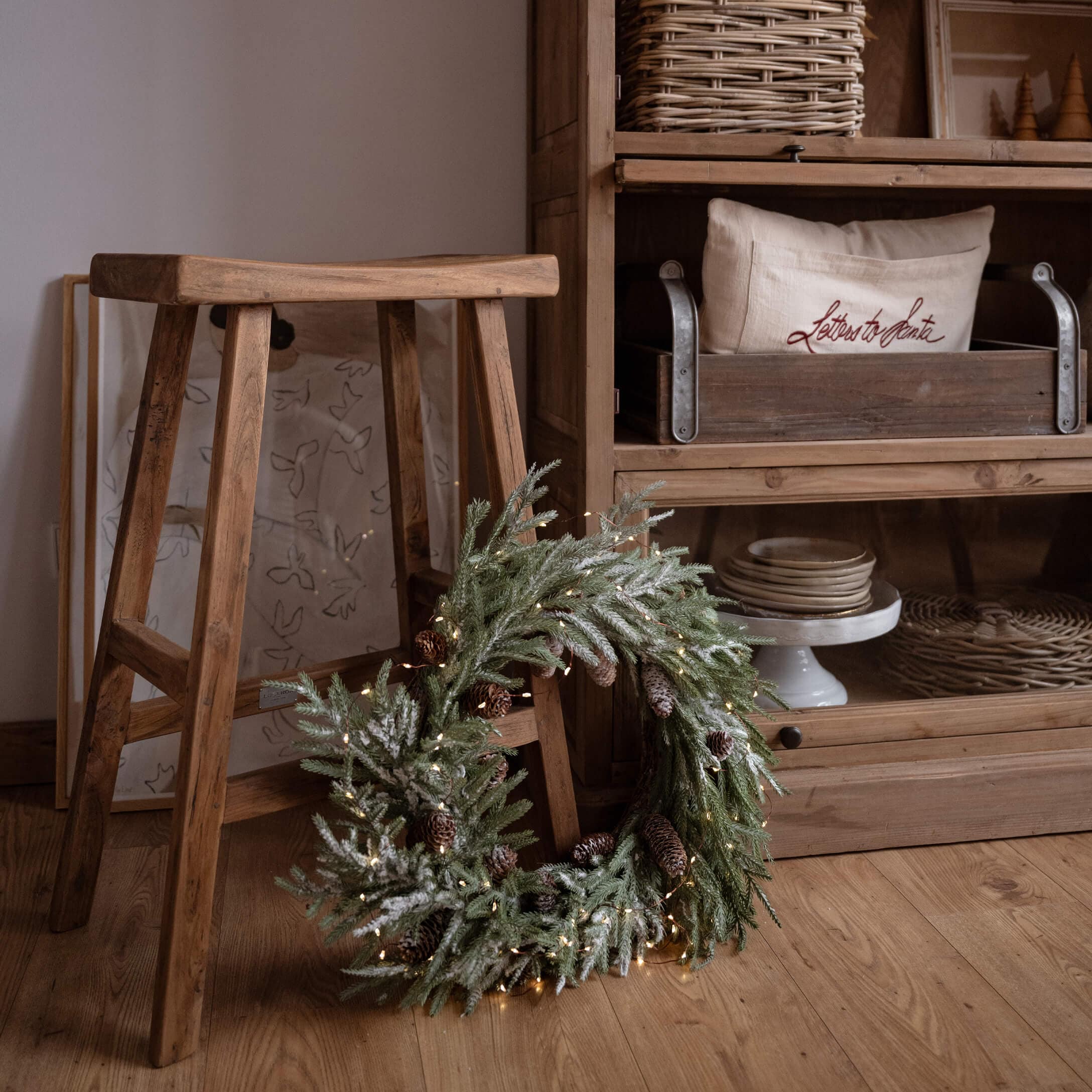 Corona natalizia decorativa con rami verdi e pigne su pavimento in legno accanto a uno sgabello