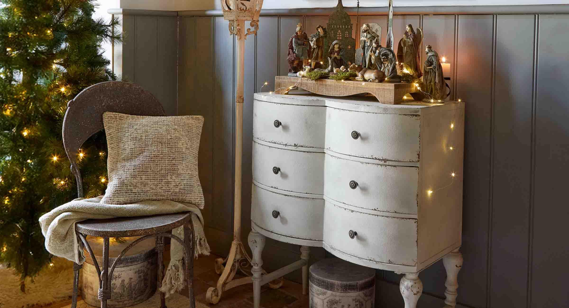 Comò bianco invecchiato con presepe in legno e decorazioni natalizie in un ambiente accogliente