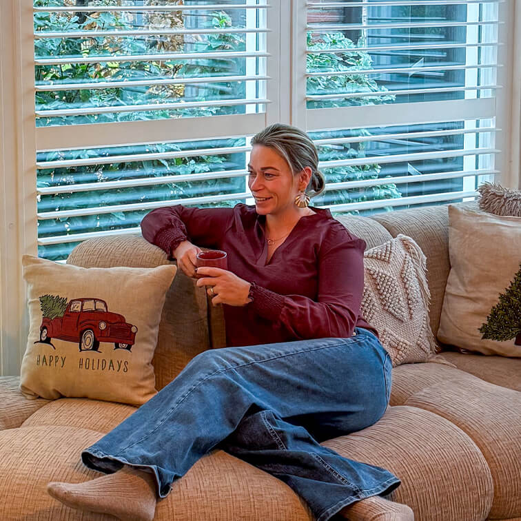 Donna con camicia bordeaux e jeans rilassata su divano beige con cuscino natalizio rosso e tazza in mano