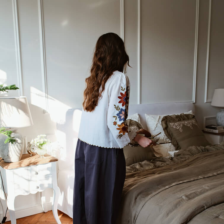 Donna con camicetta bianca ricamata a colori in camera da letto con biancheria da letto marrone e lampada sul comodino