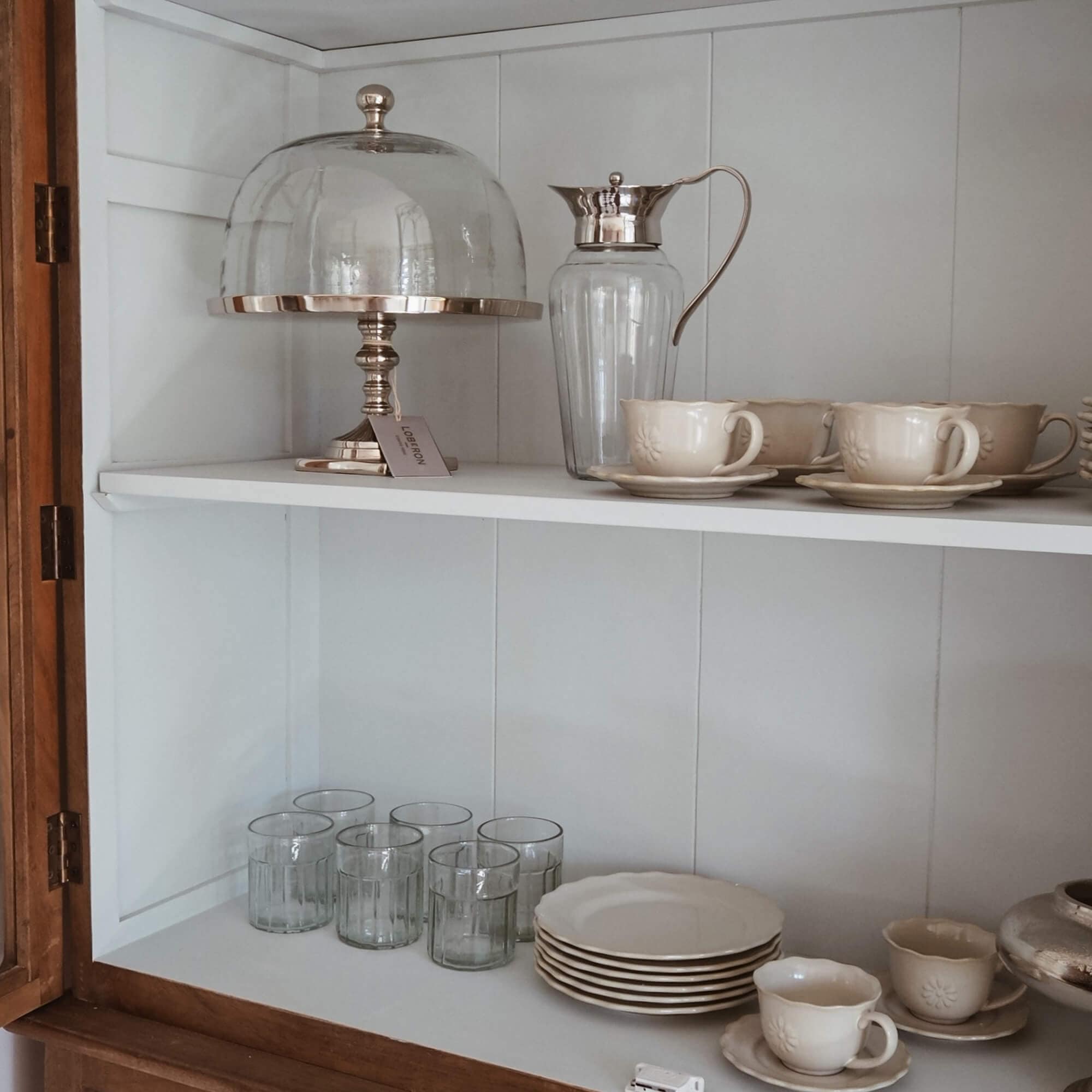 Set di tazze e piatti crema in ceramica con brocca in vetro e campana per dolci in armadio bianco