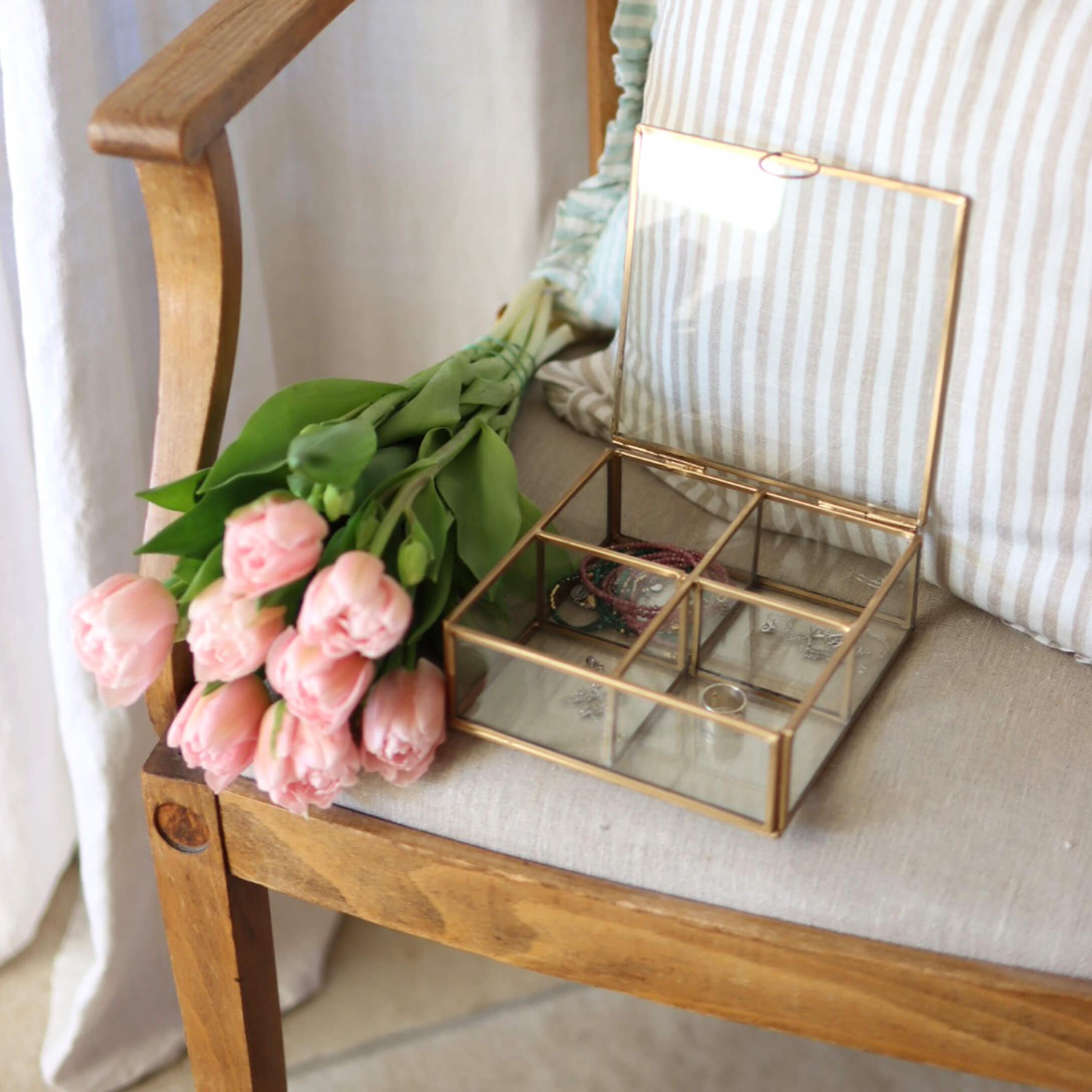 Scatola porta gioielli in vetro con cornice in ottone su sedia di legno accanto a tulipani rosa