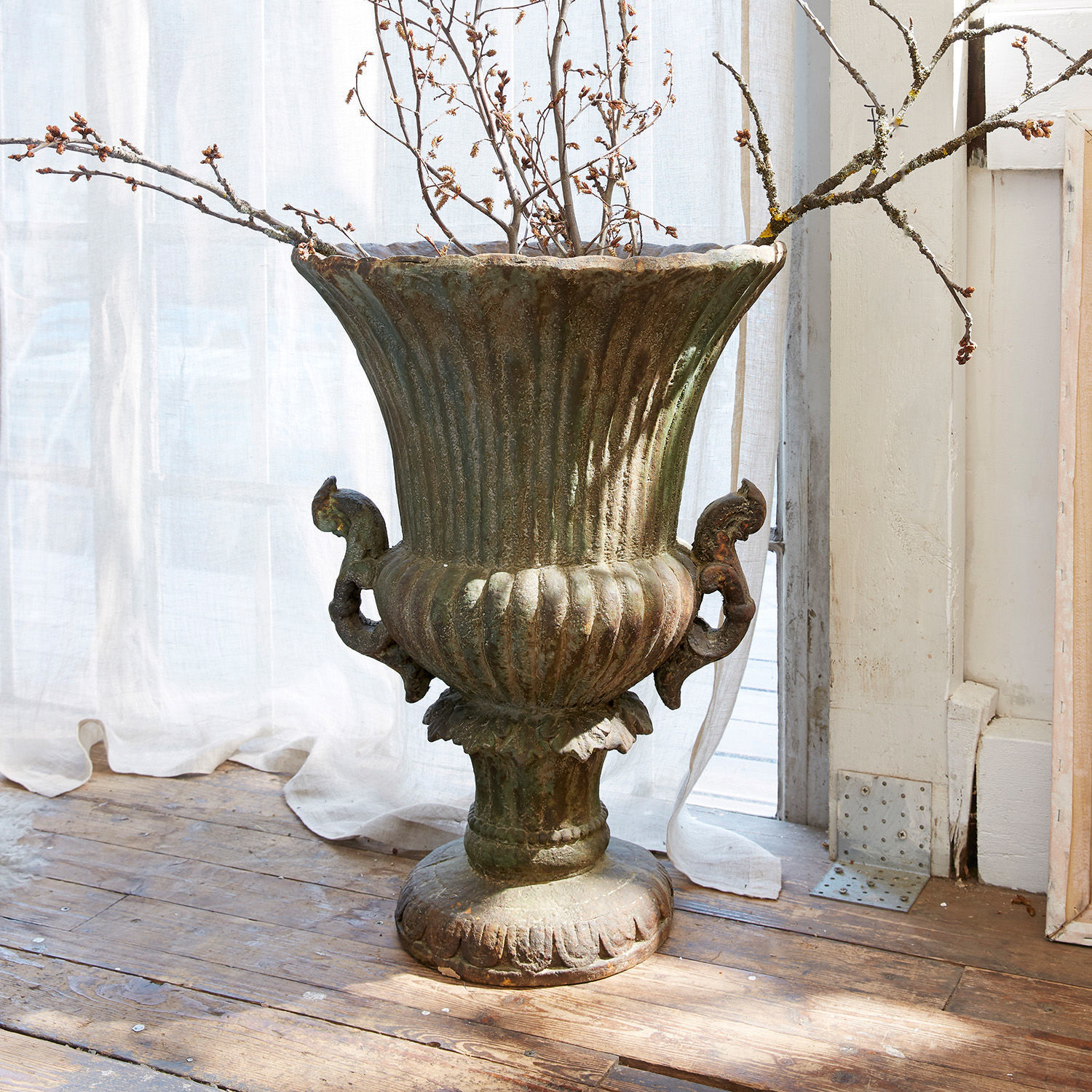 Anfora Estefanie Grande vaso decorativo in metallo verde patinato su pavimento in legno con rami secchi