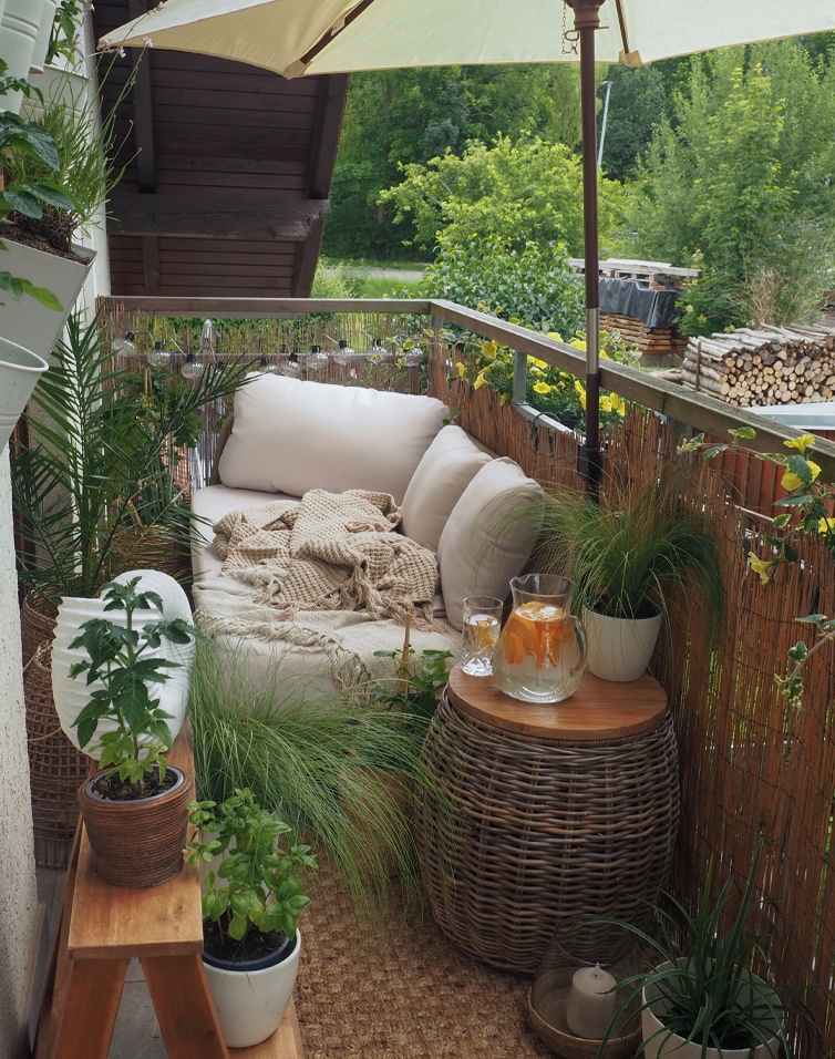 Balcone accogliente con divano beige, cuscini, tavolo in vimini, piante e ombrellone per relax