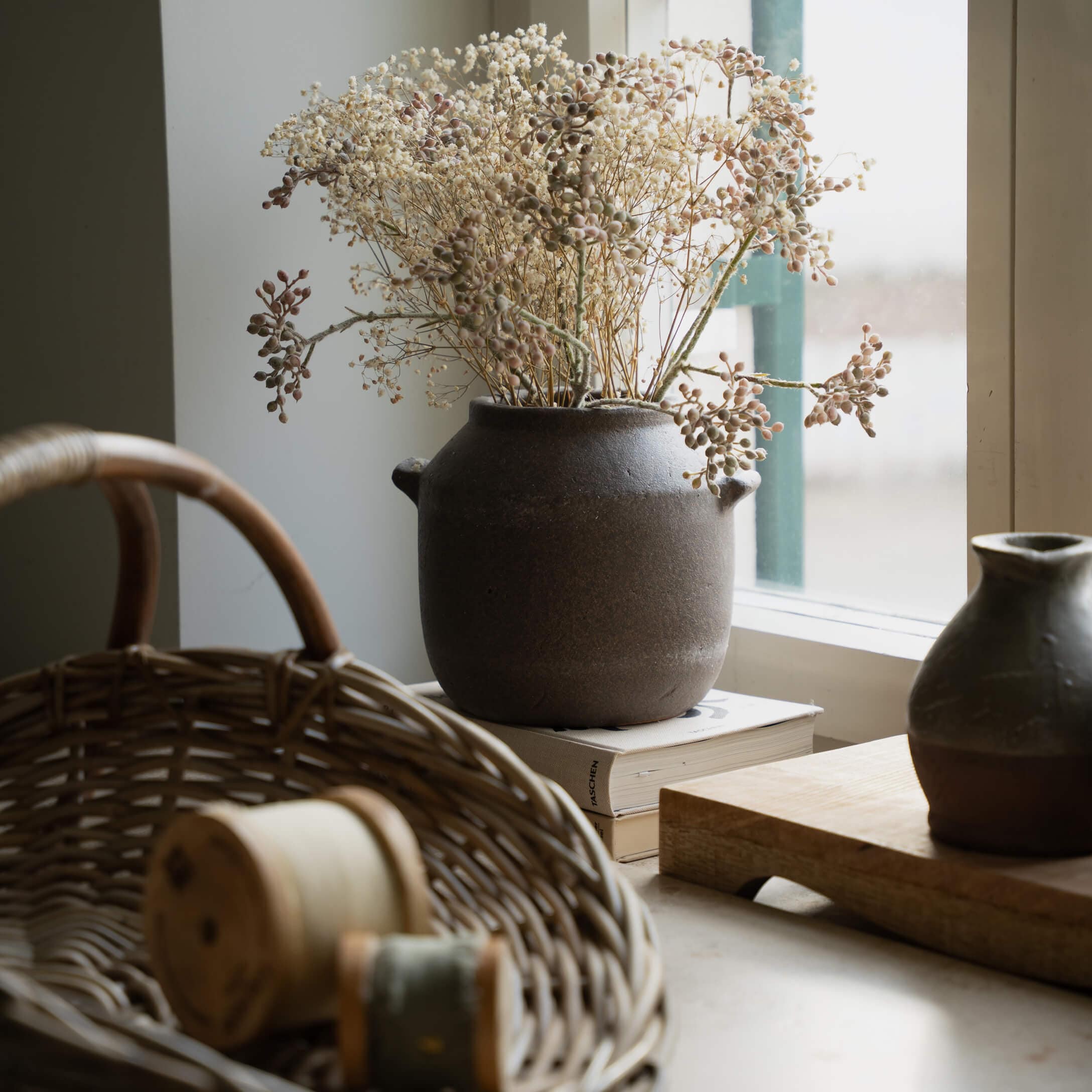 Vaso in ceramica marrone con fiori secchi sul davanzale accanto a cesto di vimini e libri