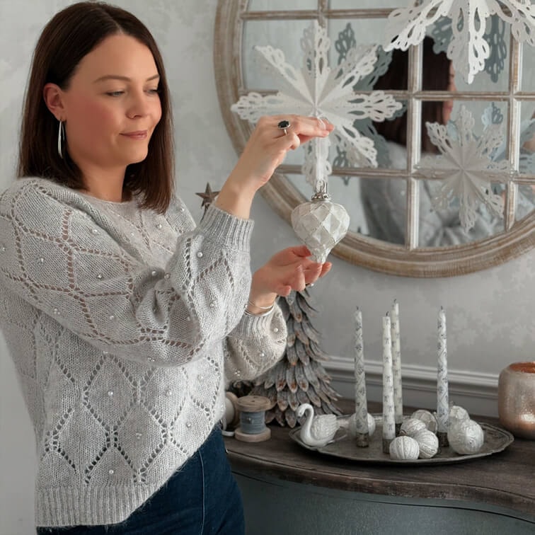 Donna con maglione grigio con perle tiene decorazione natalizia bianca in ambiente accogliente con addobbi invernali