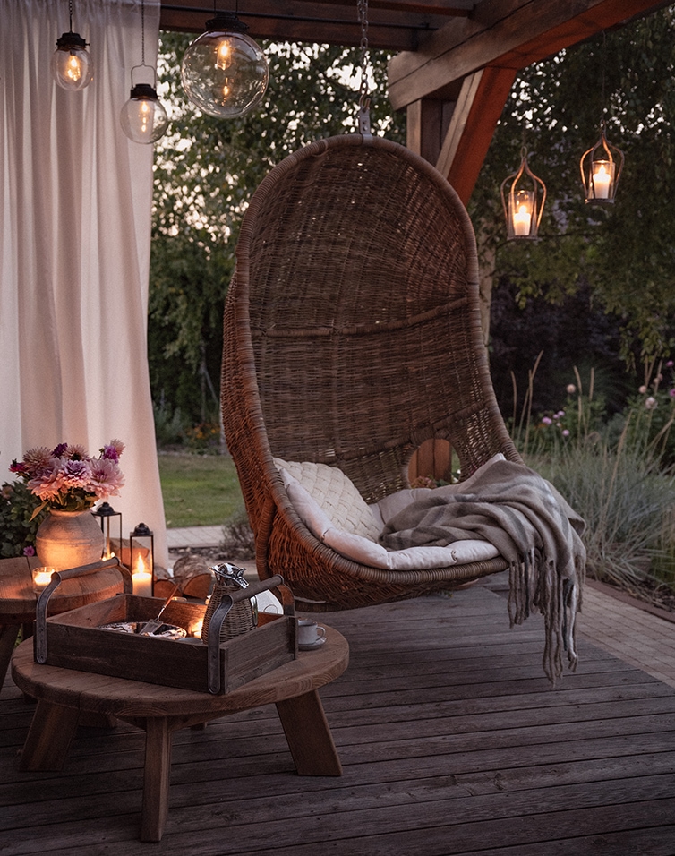 Sedia sospesa in rattan naturale su terrazza con cuscino e coperta, illuminata da lampade decorative
