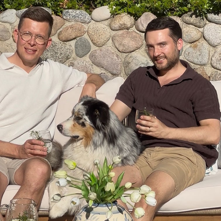 Due uomini seduti all'aperto su un divano con un cane Australian Shepherd e un vaso di tulipani bianchi.