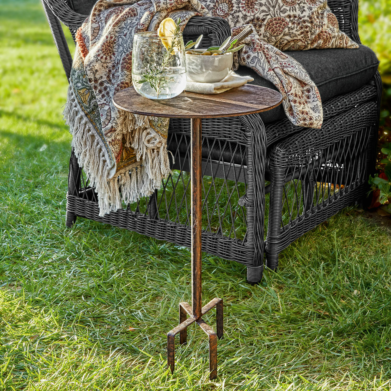 Tavolino Fermont Tavolino da giardino in metallo con patina vintage sull'erba accanto a poltrona in vimini con cuscino e coperta