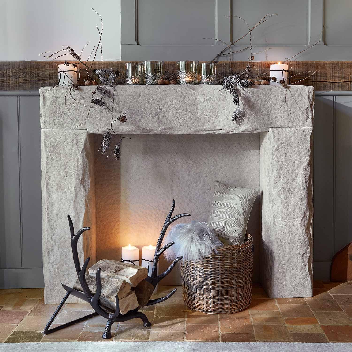 Camino in pietra bianca con decorazioni di rami e candele, cesta in vimini con cuscino e coperta di pelliccia
