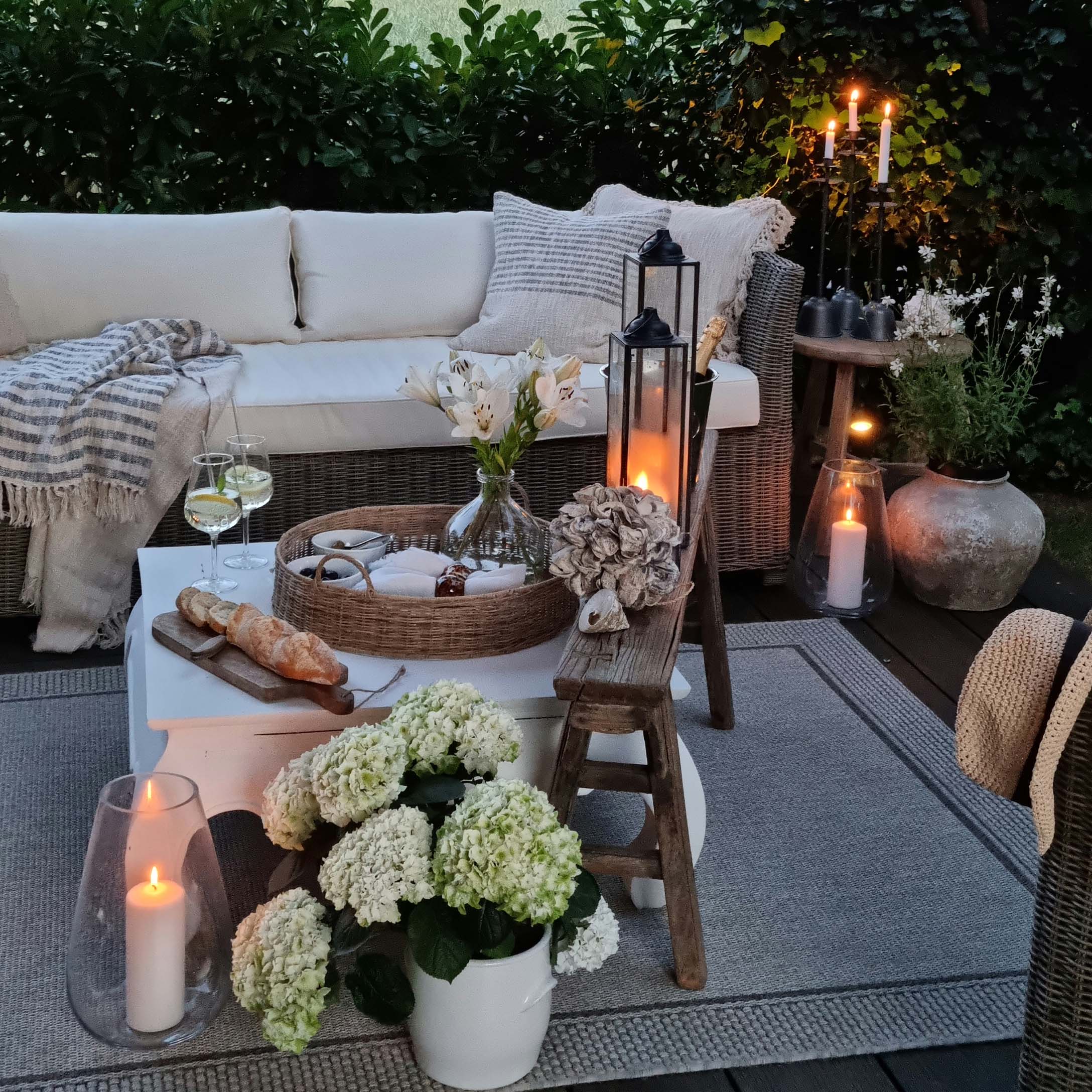 Angolo giardino accogliente con divano in rattan, cuscini bianchi e candele su terrazza in legno