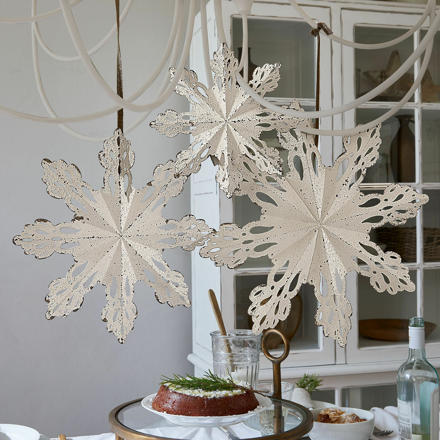 Set di 3 decorazioni da parete Lirvaya Fiocchi di neve decorativi in metallo color crema appesi in ambiente luminoso con torta sul tavolo