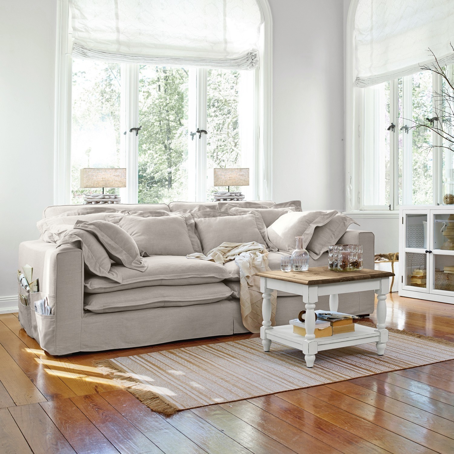 Divano beige in tessuto con cuscini e tavolino bianco su pavimento in legno in soggiorno luminoso