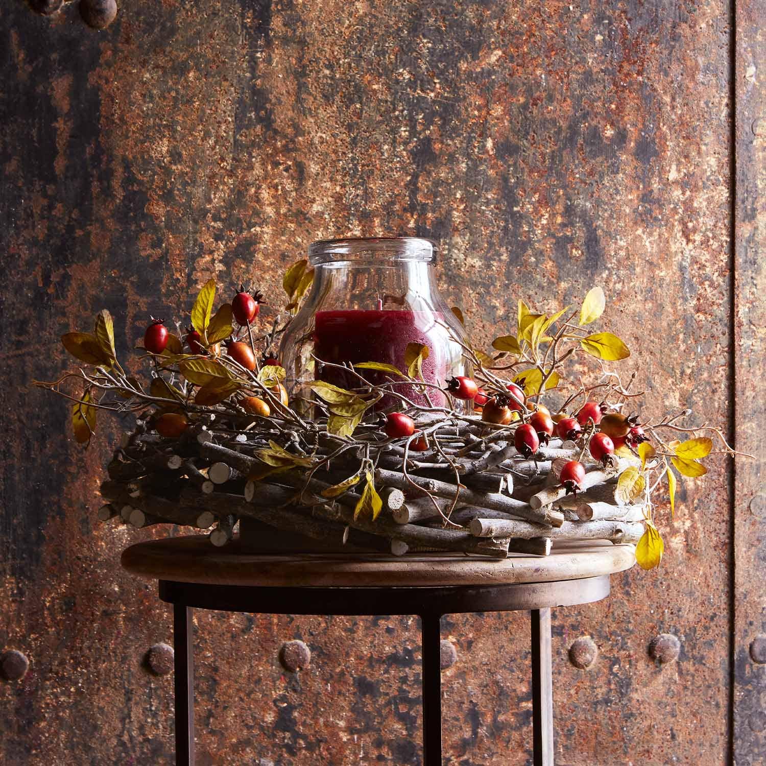 Corona decorativa di rami con bacche rosse e candela rossa in vetro su tavolo in legno