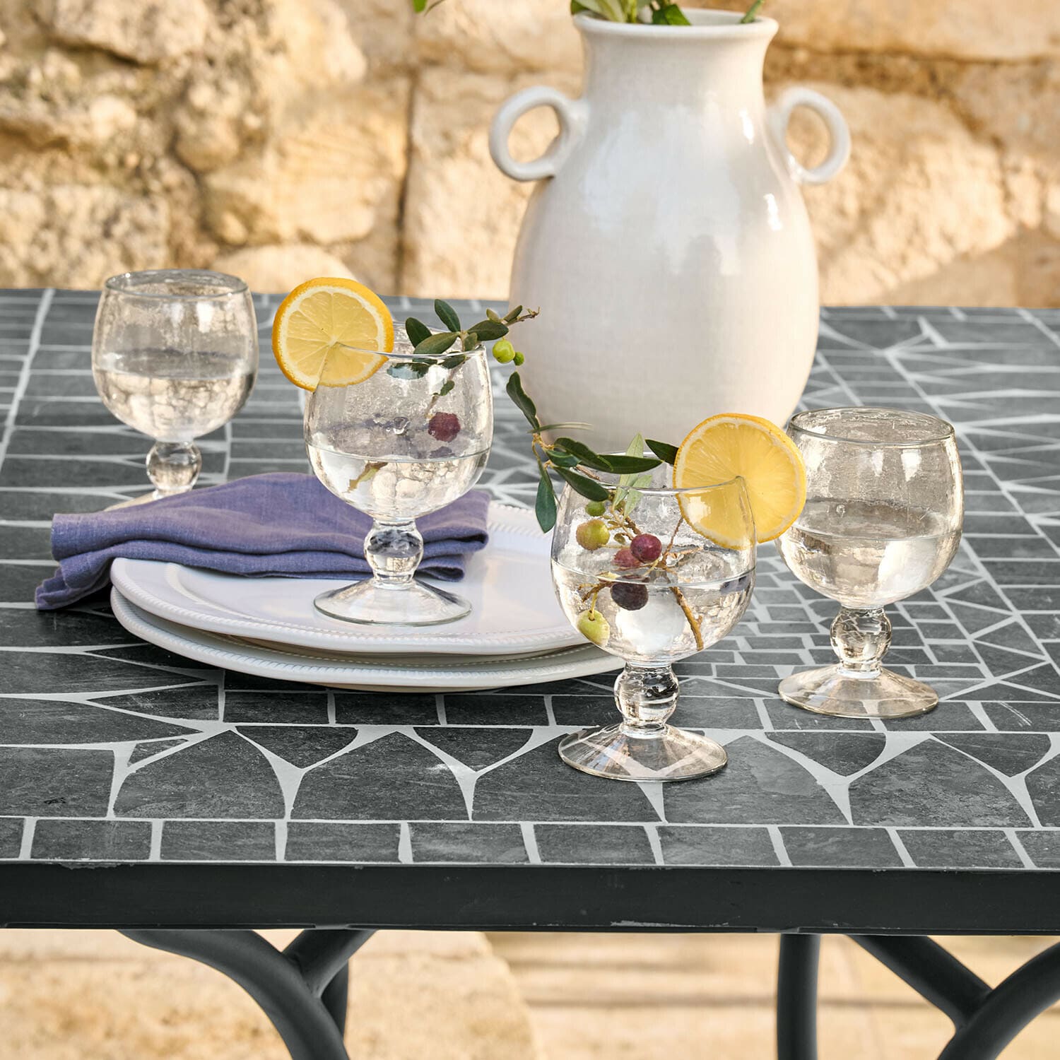 Set di 4 bicchieri Vinoros Bicchieri di vetro con acqua e fette di limone su tavolo nero a mosaico, vaso in ceramica bianca sullo sfondo