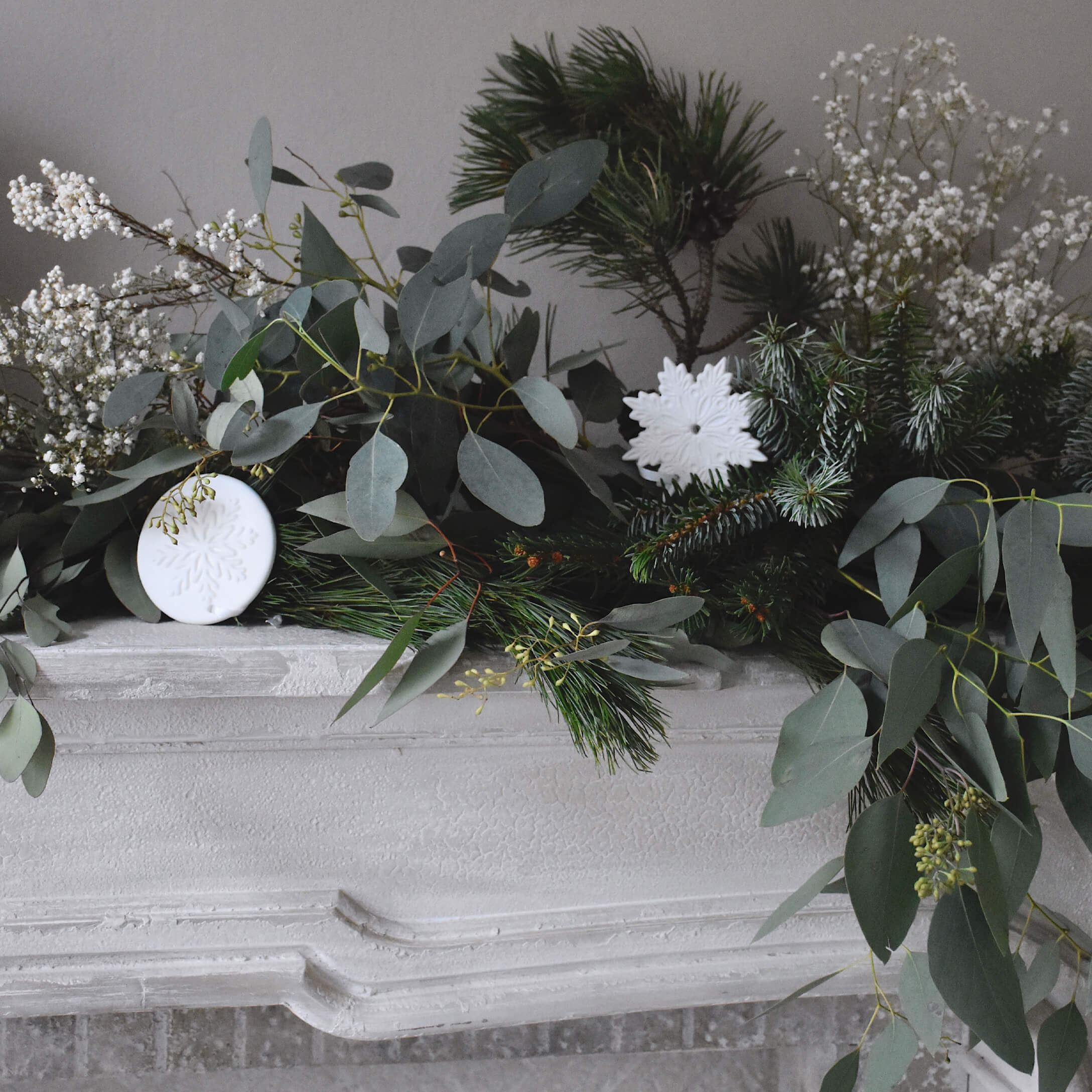 Corona decorativa con rami verdi e ornamenti bianchi a forma di fiocco di neve su camino in pietra bianca