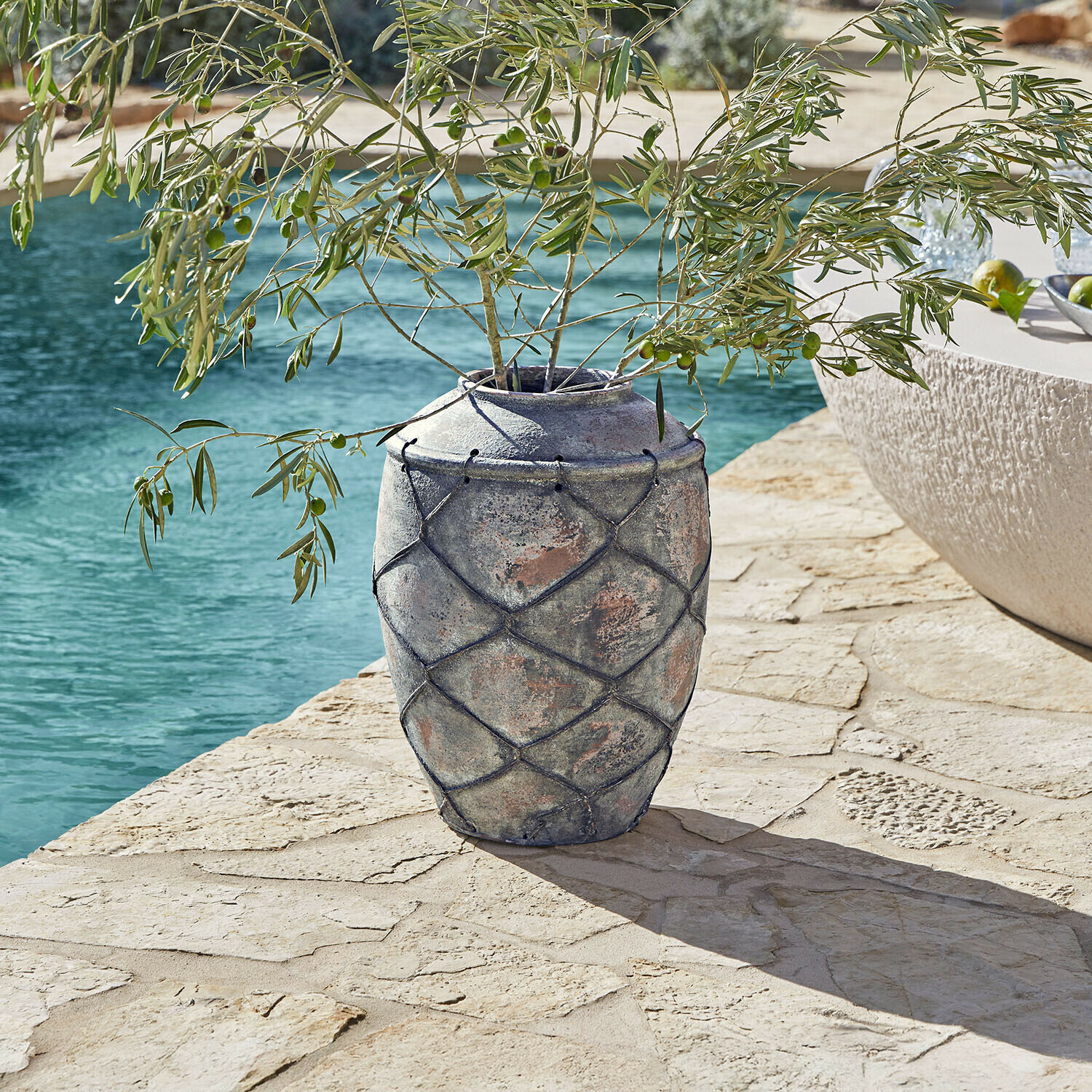 Grande vaso in ceramica grigia con rete decorativa su terrazza in pietra vicino a piscina con rami di ulivo