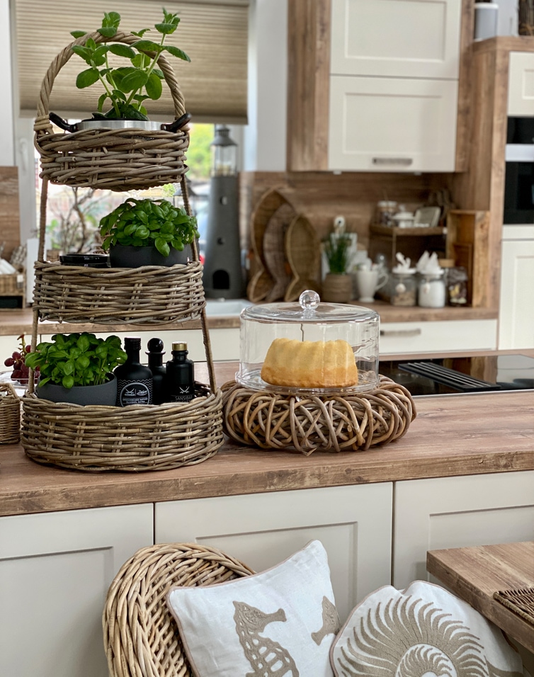 Portaoggetti a tre livelli in vimini con piante verdi e campana di vetro per dolci su piano cucina in legno