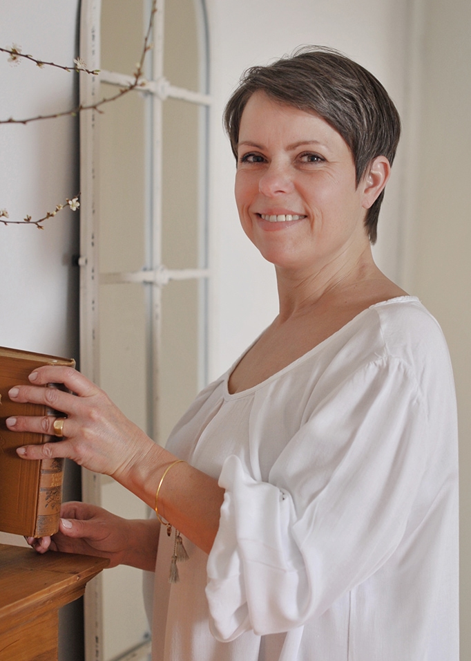 Donna con camicetta bianca di cotone che tiene un vecchio libro vicino a una mensola di legno in una stanza luminosa