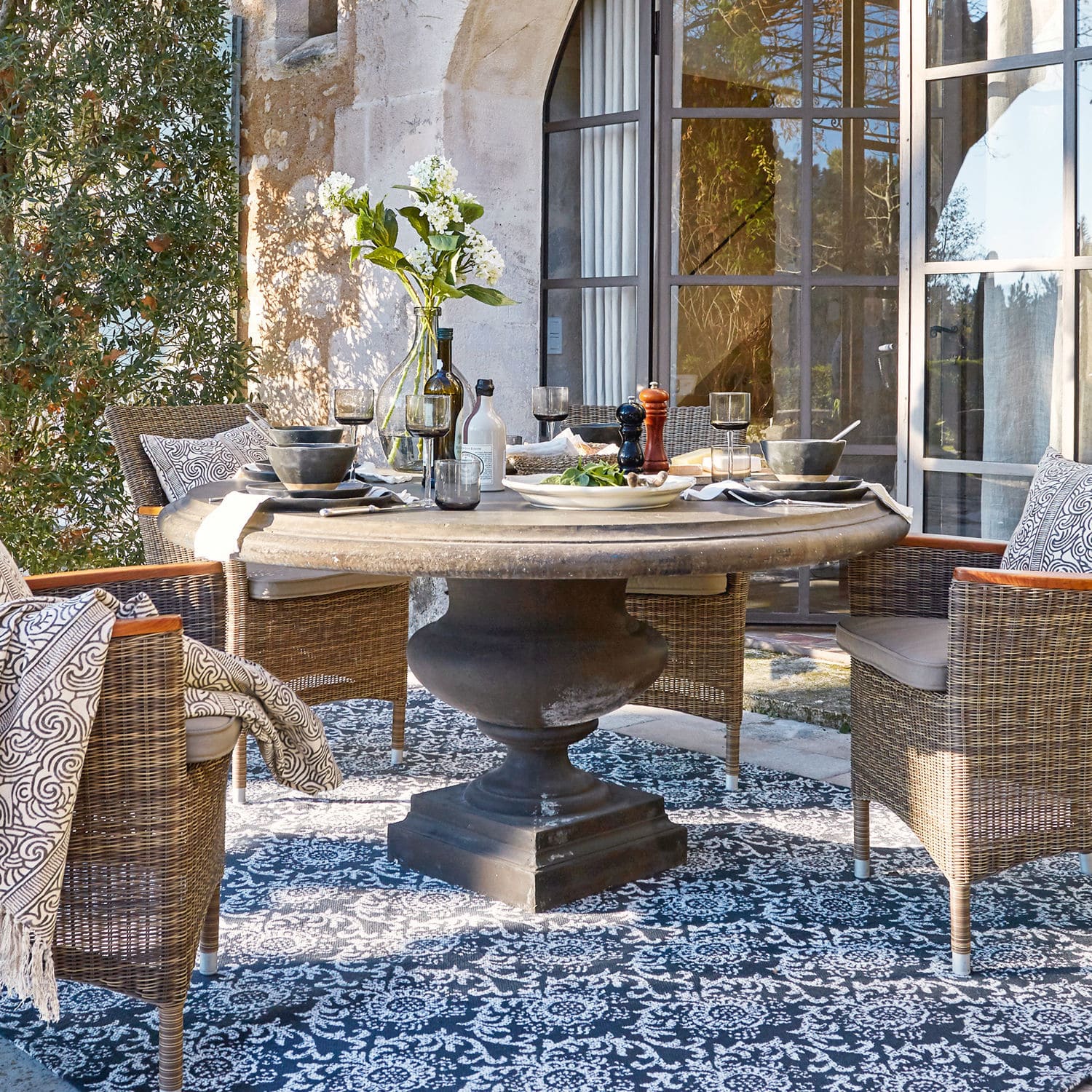 Tavolo Monterey Tavolo rotondo in pietra con base nera e sedie in vimini su terrazza con tappeto decorativo